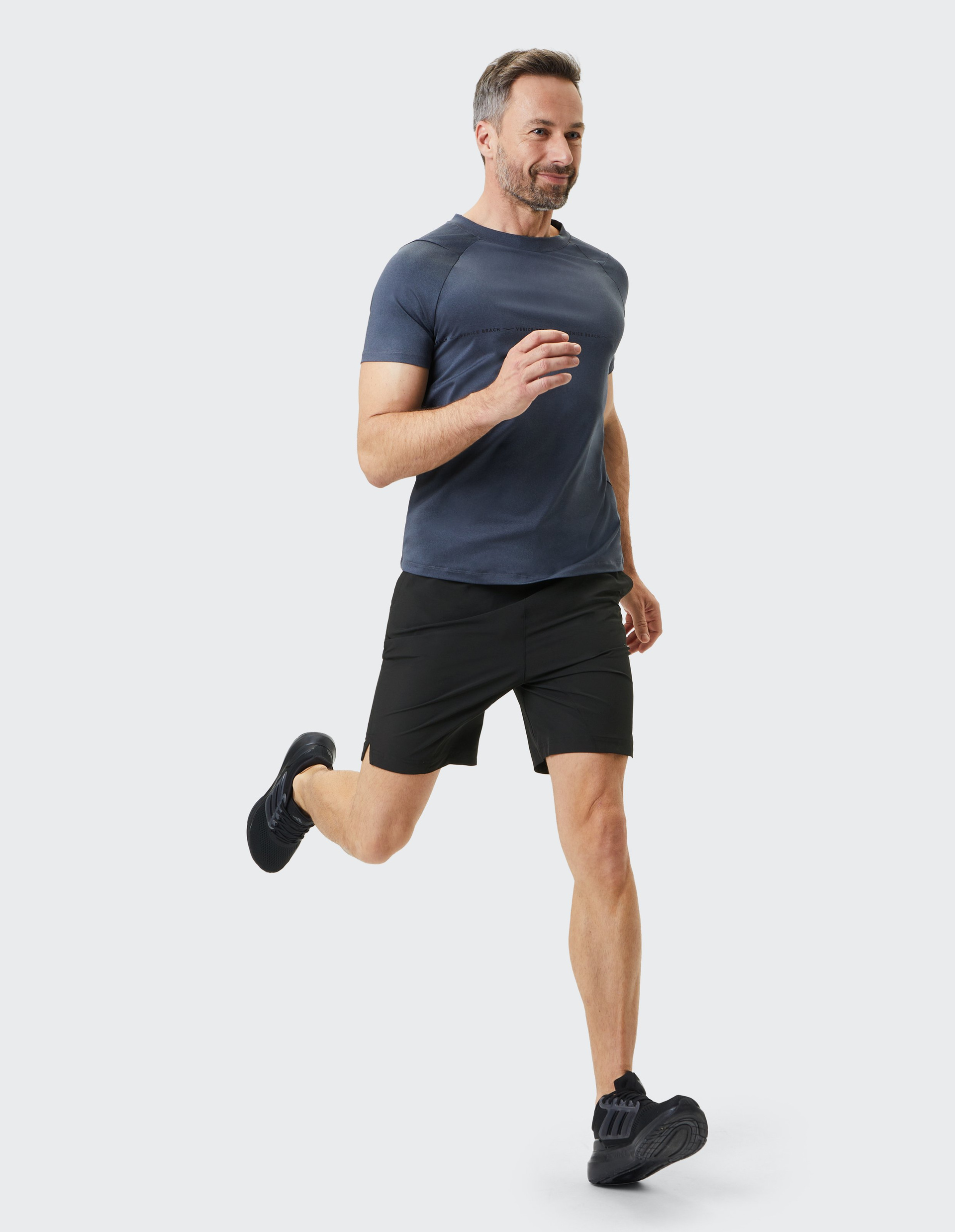 Mann in sportlicher Kleidung joggt, trägt ein dunkelblaues T-Shirt, schwarze Shorts und Schuhe, in dynamischer Laufbewegung vor hellem Hintergrund.