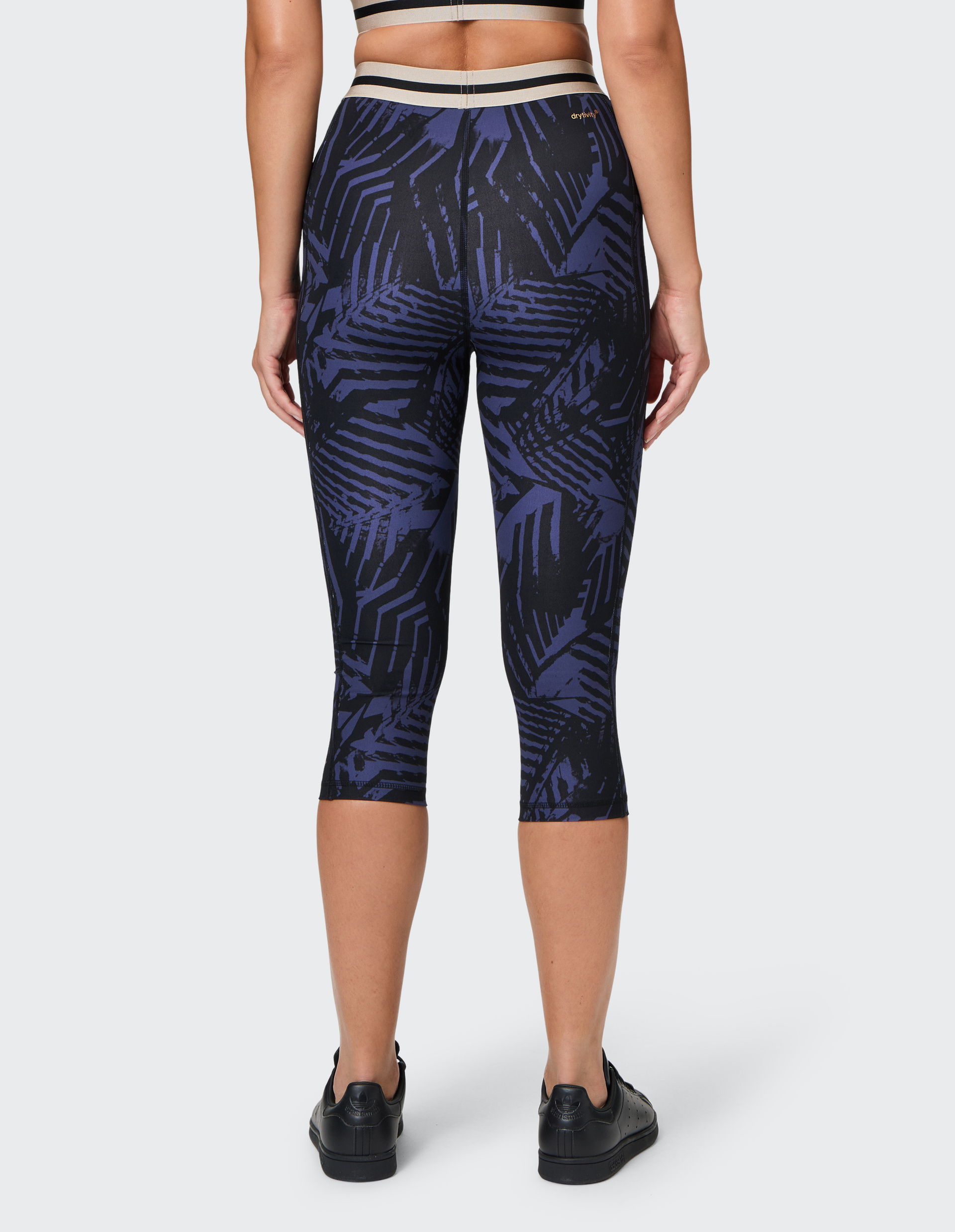 Rückansicht einer Person mit schwarzen Sneakers und gemusterten, knielangen Leggings mit blau-schwarzem Design vor neutralem Hintergrund.