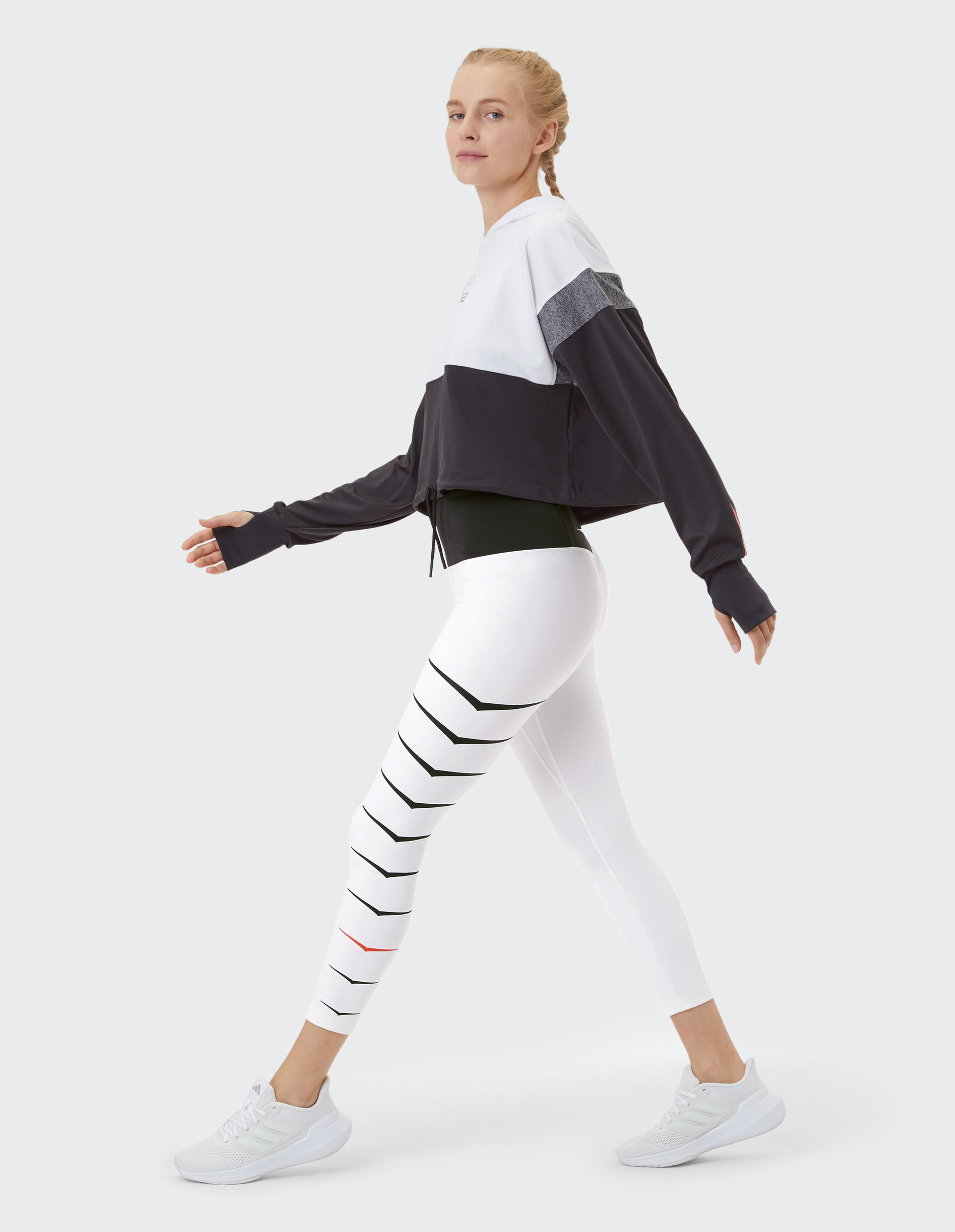 Frau in schwarz-weißem Sportoutfit mit gestreifter Leggings und Sneakers geht auf hellem Hintergrund seitwärts.