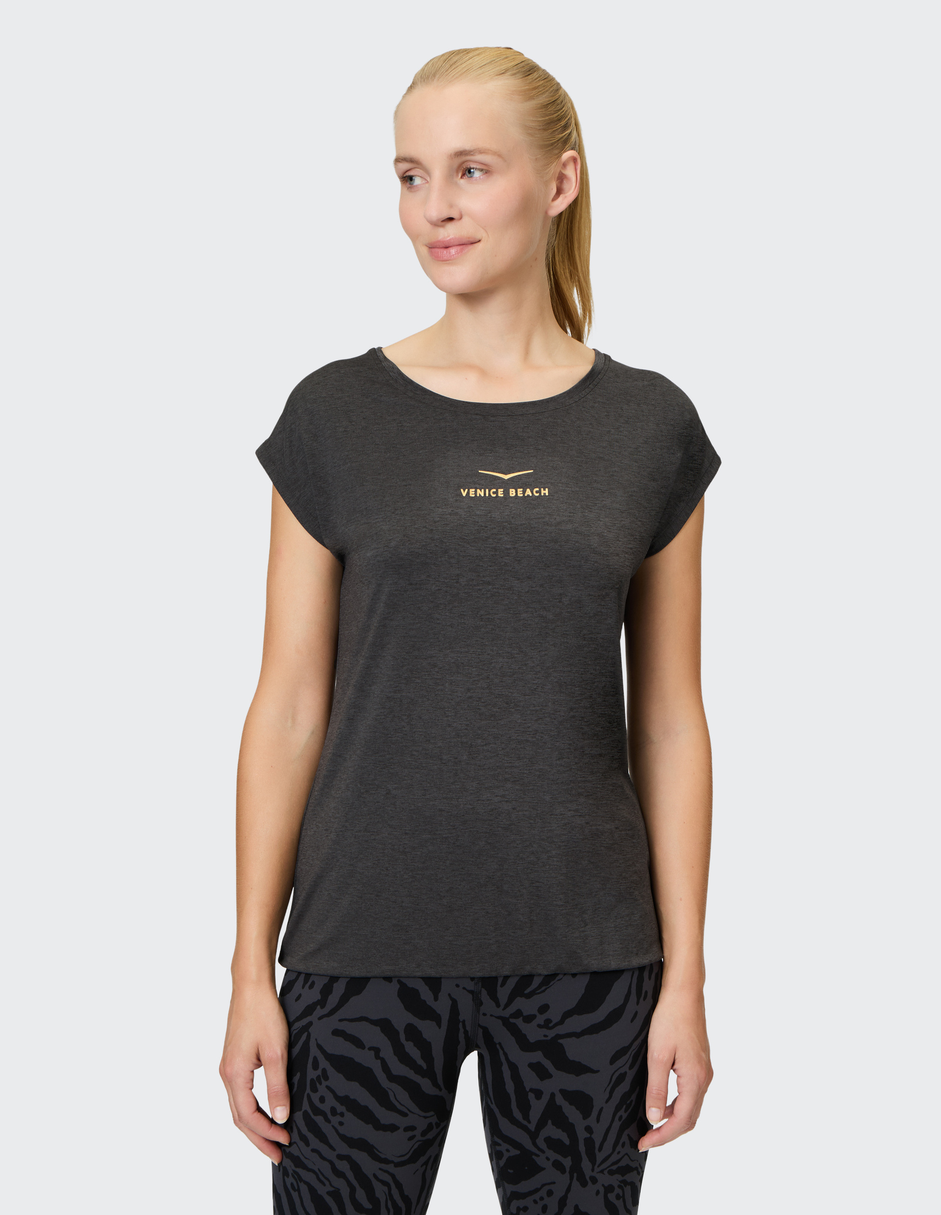 Frau trägt ein graues T-Shirt mit "Venice Beach"-Schrift und gemusterter Leggings, schaut seitlich, blonder Pferdeschwanz, hellgrauer Hintergrund.
