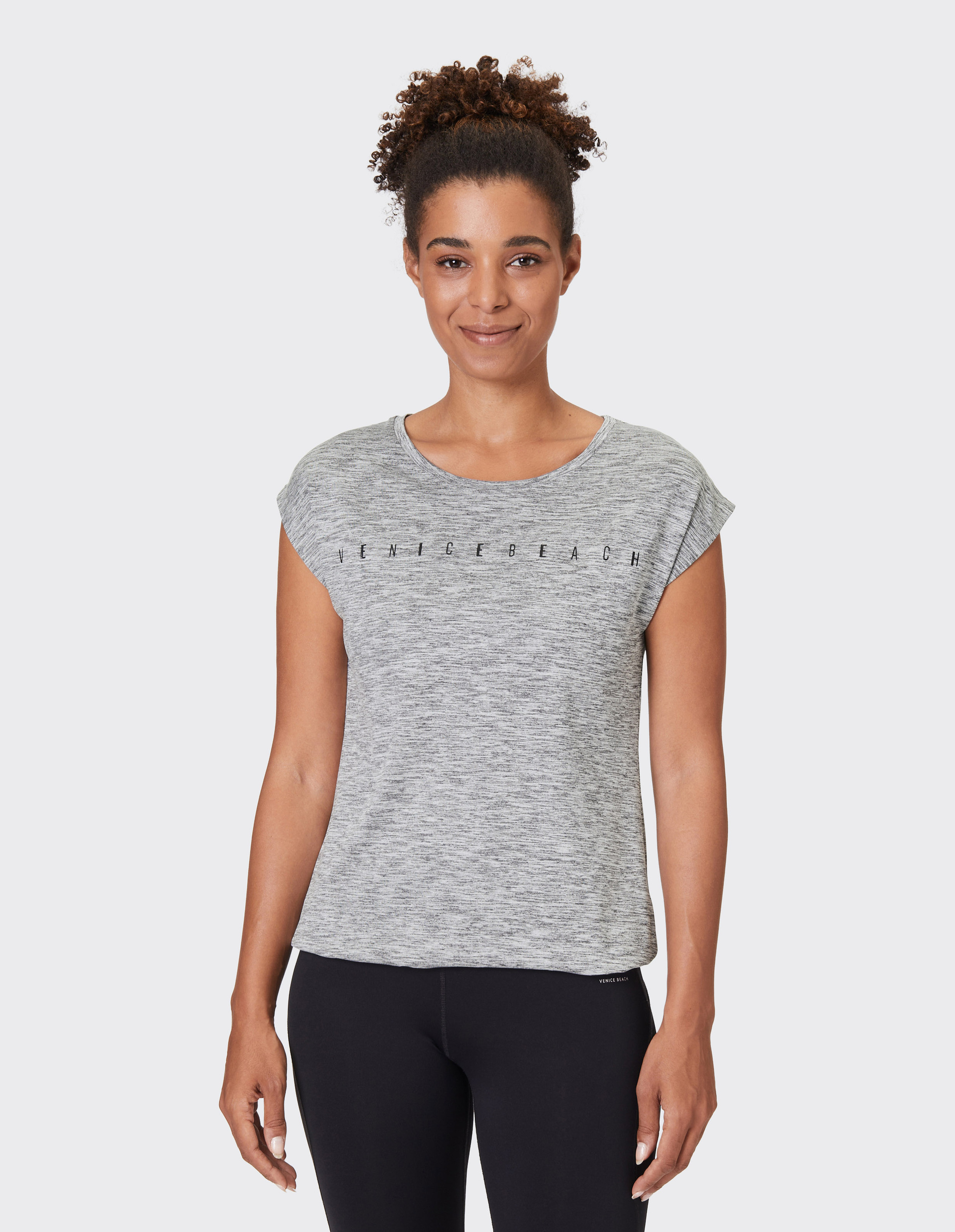 Frau mit lockigem Haar trägt ein graues T-Shirt mit der Aufschrift "Venice Beach" und schwarze Leggings, lächelnd vor neutralem Hintergrund.