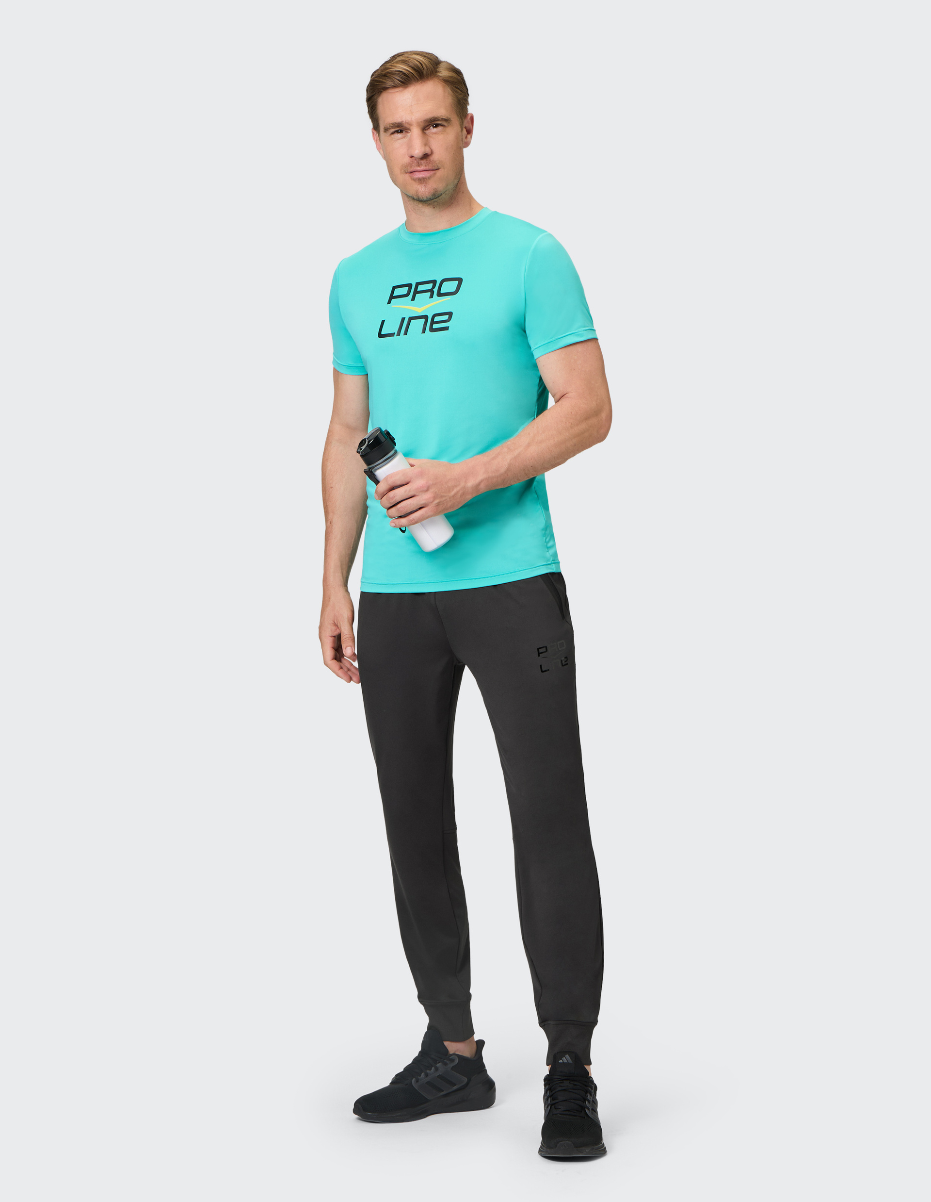 Mann in türkisfarbenem "Pro Line"-T-Shirt und schwarzer Jogginghose hält eine Wasserflasche und trägt schwarze Turnschuhe.