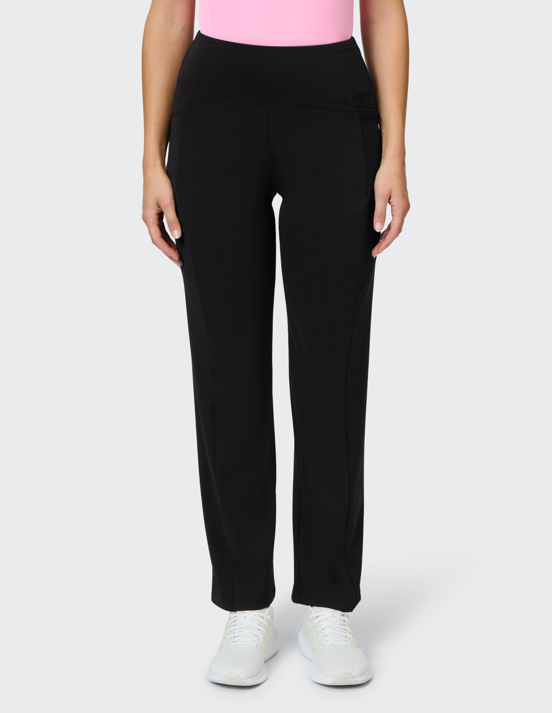 Eine Person trägt eine schwarze Hose und weiße Turnschuhe, kombiniert mit einem rosa Oberteil, vor grauem Hintergrund.