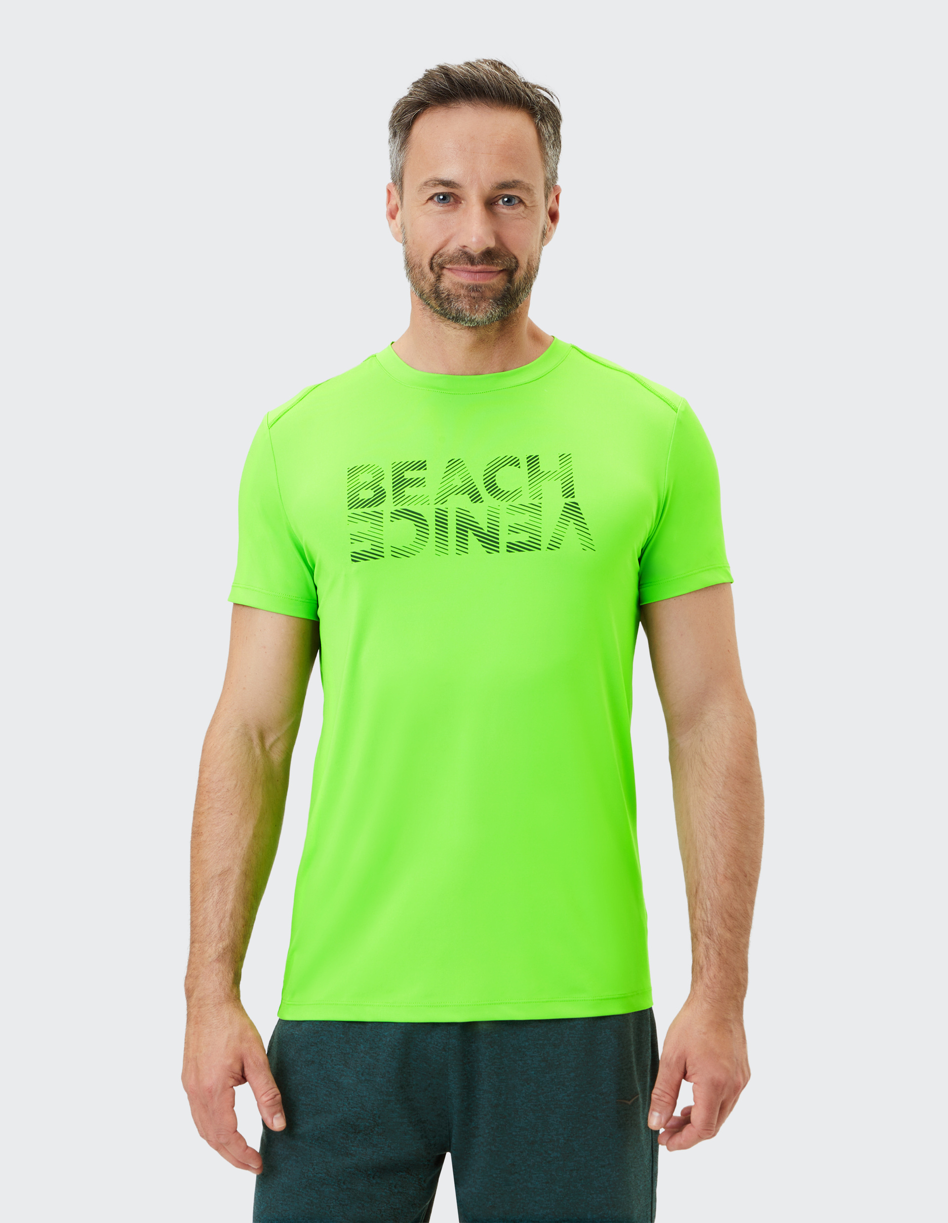 Mann in neon-grünem T-Shirt mit dem Aufdruck "BEACH VIBES", lächelt leicht, trägt dunkle Hose, vor hellem Hintergrund.
