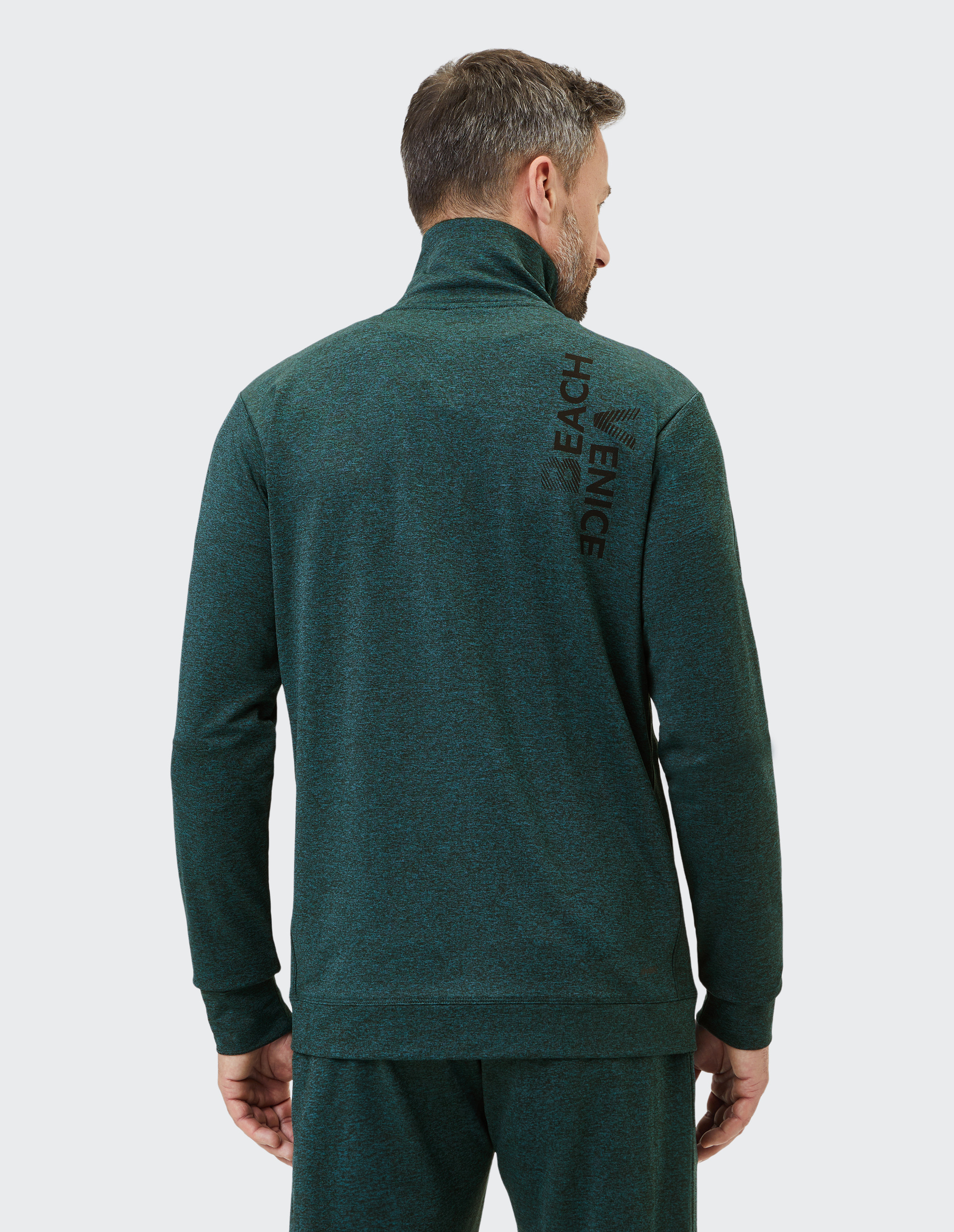 Mann in dunkelgrünem Trainingsanzug mit hohem Kragen, Rückansicht; "VENICE BEACH" in schwarzer Schrift auf der Schulter.