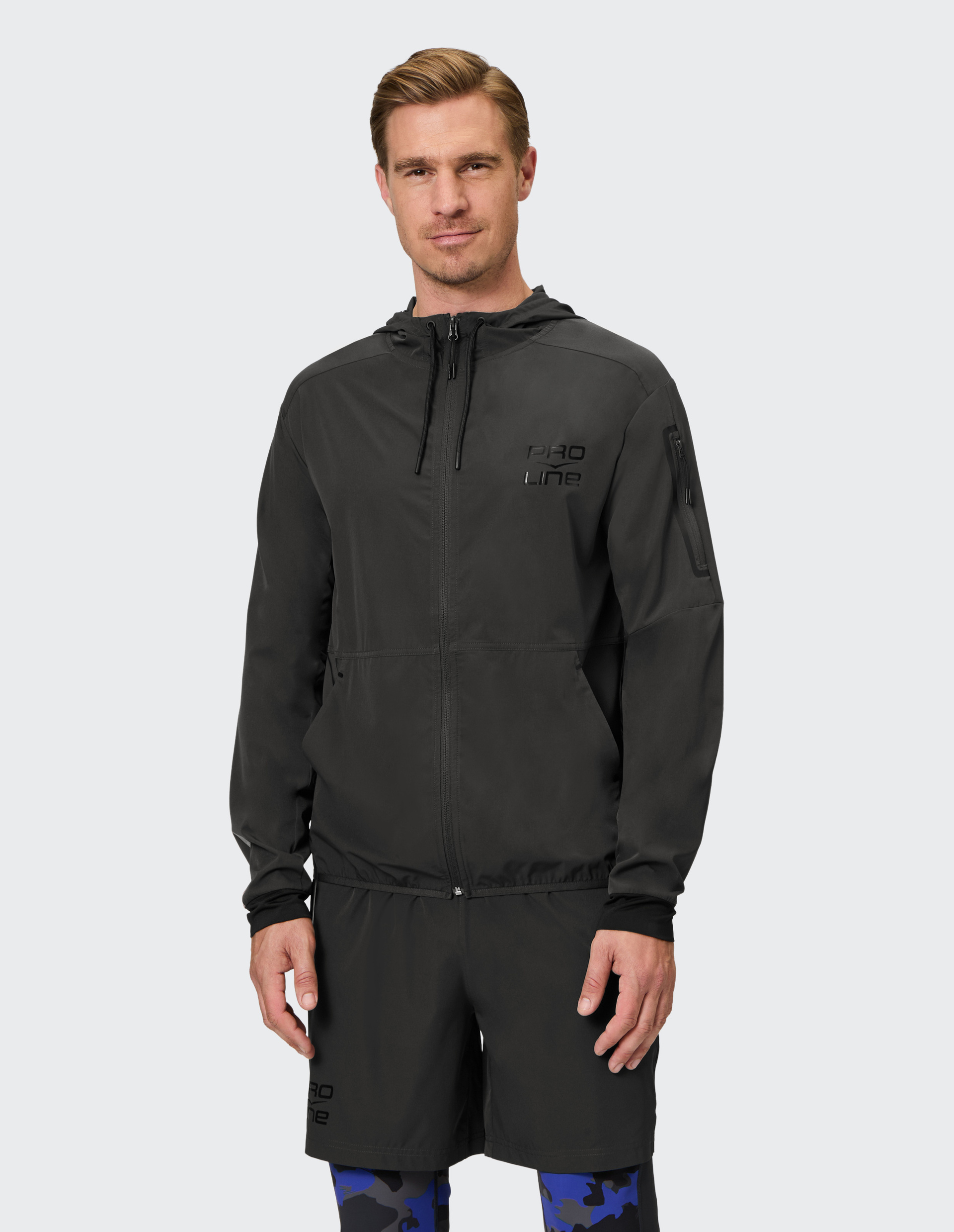 Ein Mann trägt eine schwarze Sportjacke mit Kapuze und passenden Shorts, ergänzt durch eine Hose mit blau-schwarzem Tarnmuster.