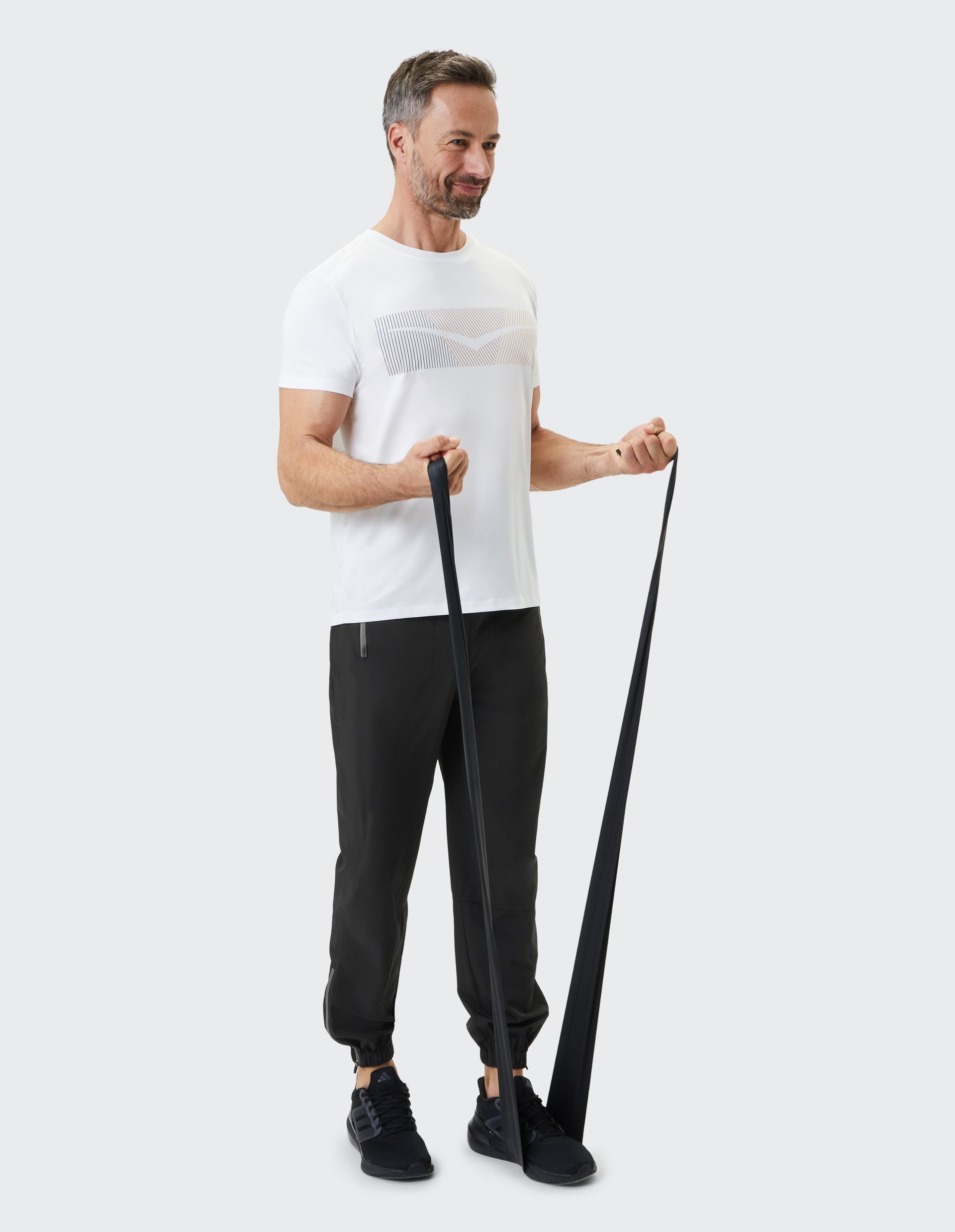Ein Mann in Sportkleidung trainiert mit einem schwarzen Fitnessband, trägt ein weißes T-Shirt und schwarze Jogginghosen.