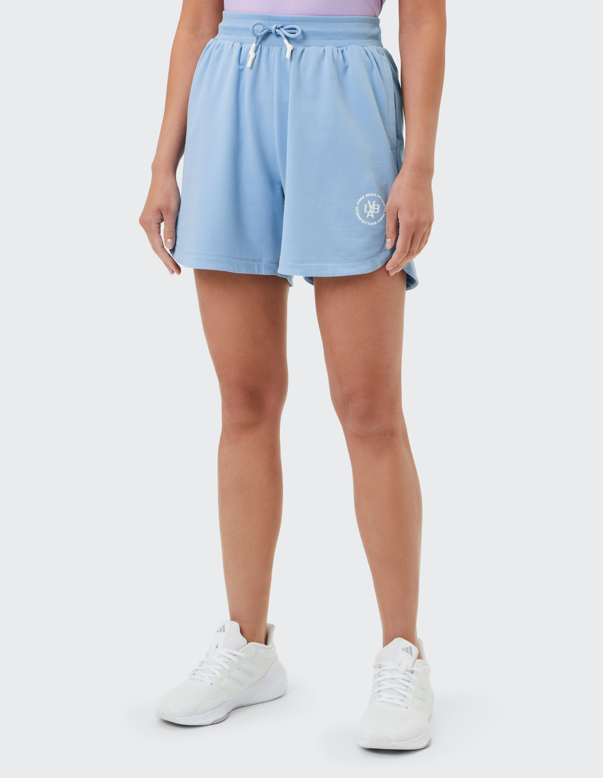 Person trägt hellblaue Shorts mit Kordelzug und weißem Logo sowie weiße Sneakers und ein lila Oberteil, vor weißem Hintergrund.