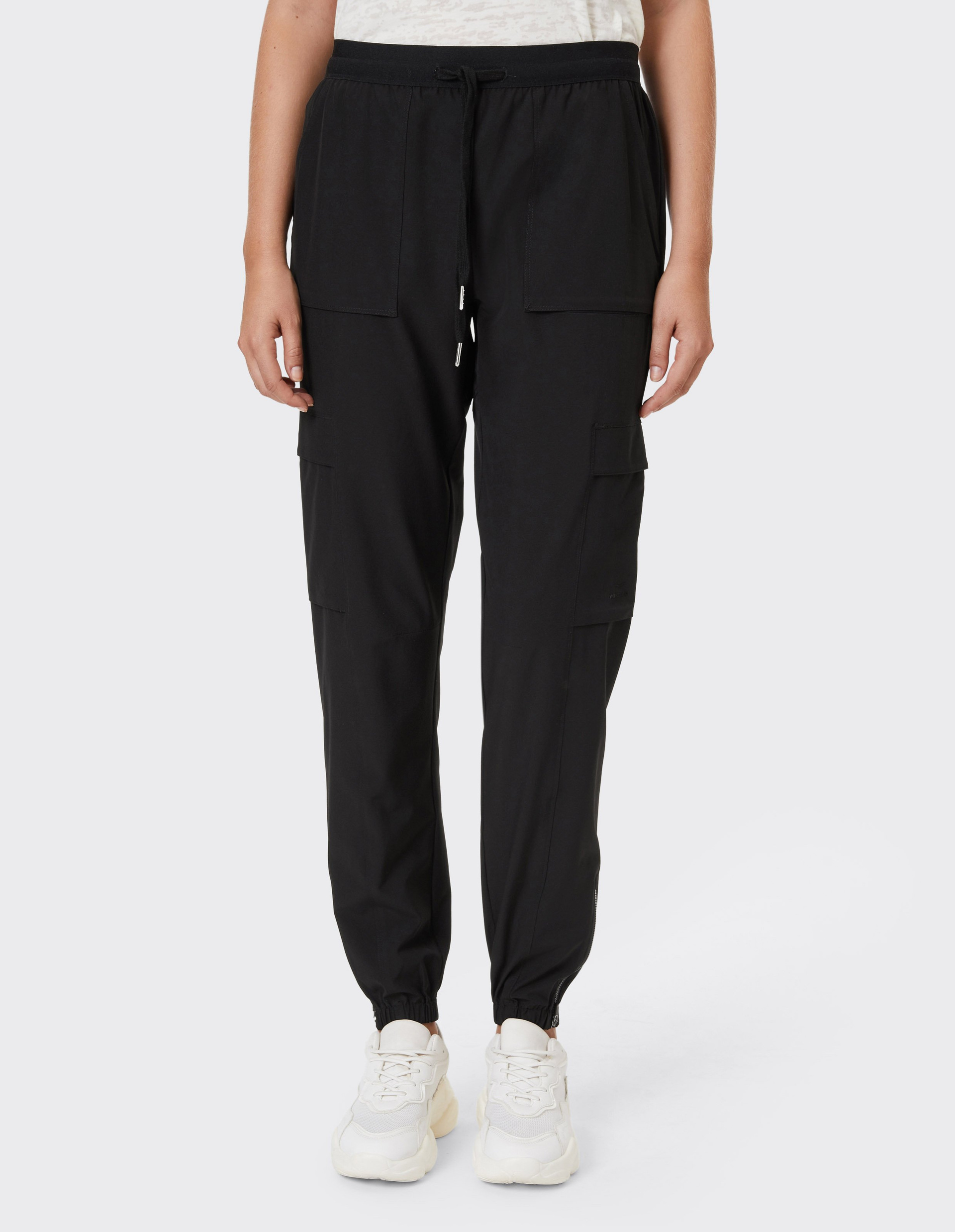 Person trägt schwarze Jogginghose mit Kordelzug und großen Taschen, dazu weiße Sneakers und ein helles Shirt, vor grauem Hintergrund.