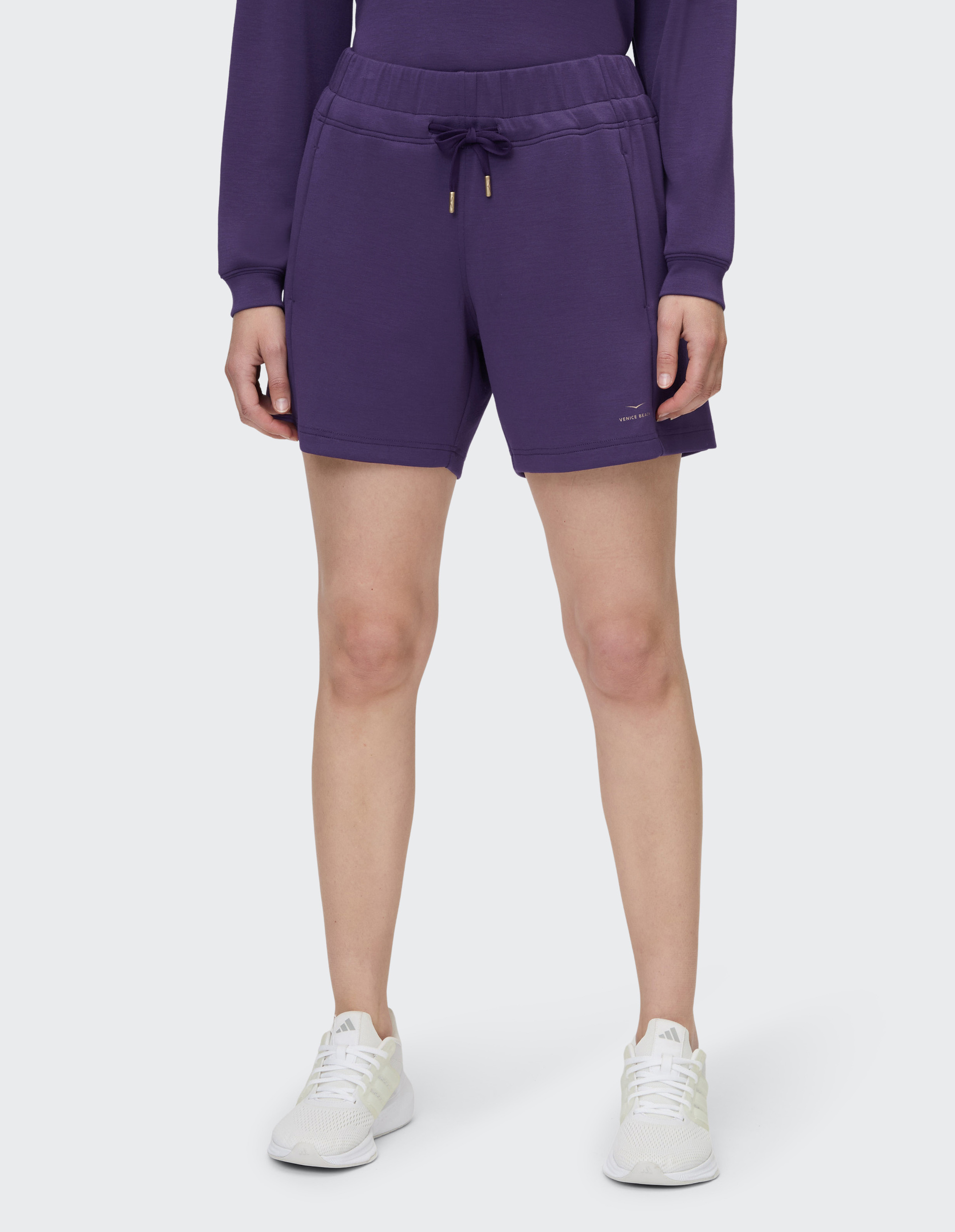 Frau in lila Kapuzenpullover und Shorts, trägt weiße Turnschuhe, steht aufrecht vor einem weißen Hintergrund.