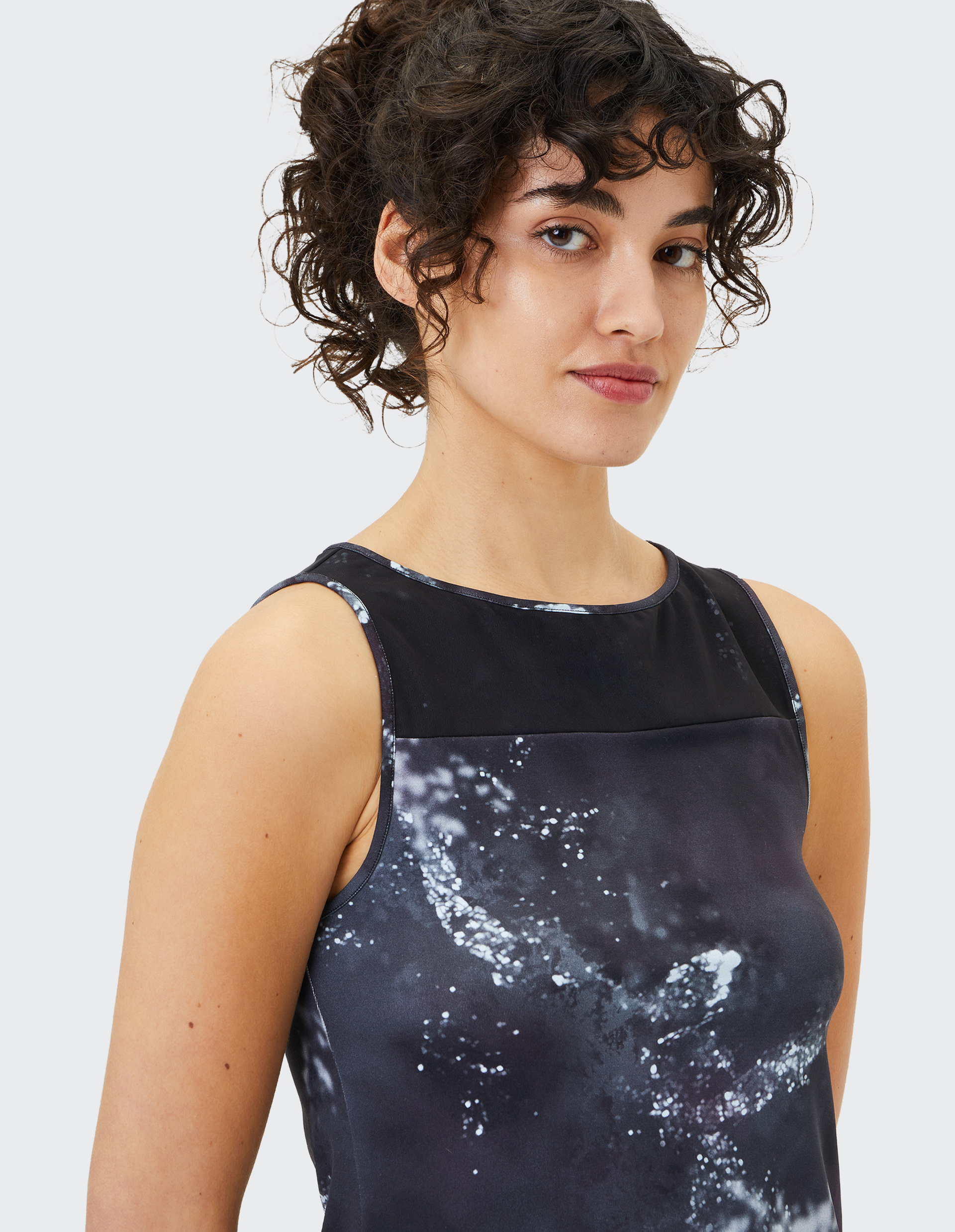 Eine Frau mit lockigem Haar trägt ein ärmelloses, dunkel gemustertes Oberteil und blickt mit leichtem Lächeln in die Kamera.