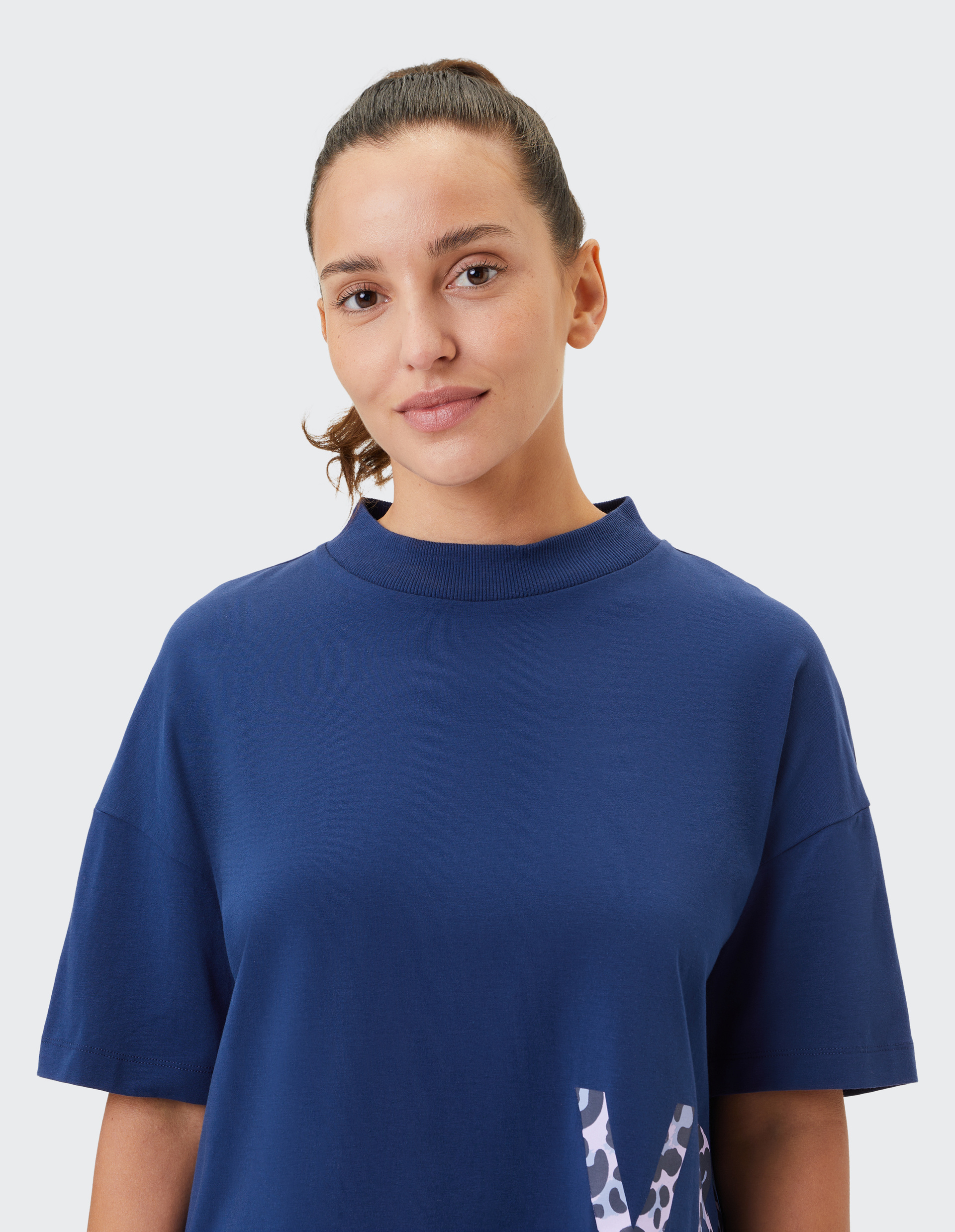 Frau in blauem T-Shirt mit Pferdeschwanz und neutralem Hintergrund, der obere Teil des Shirts zeigt ein abstraktes Muster.