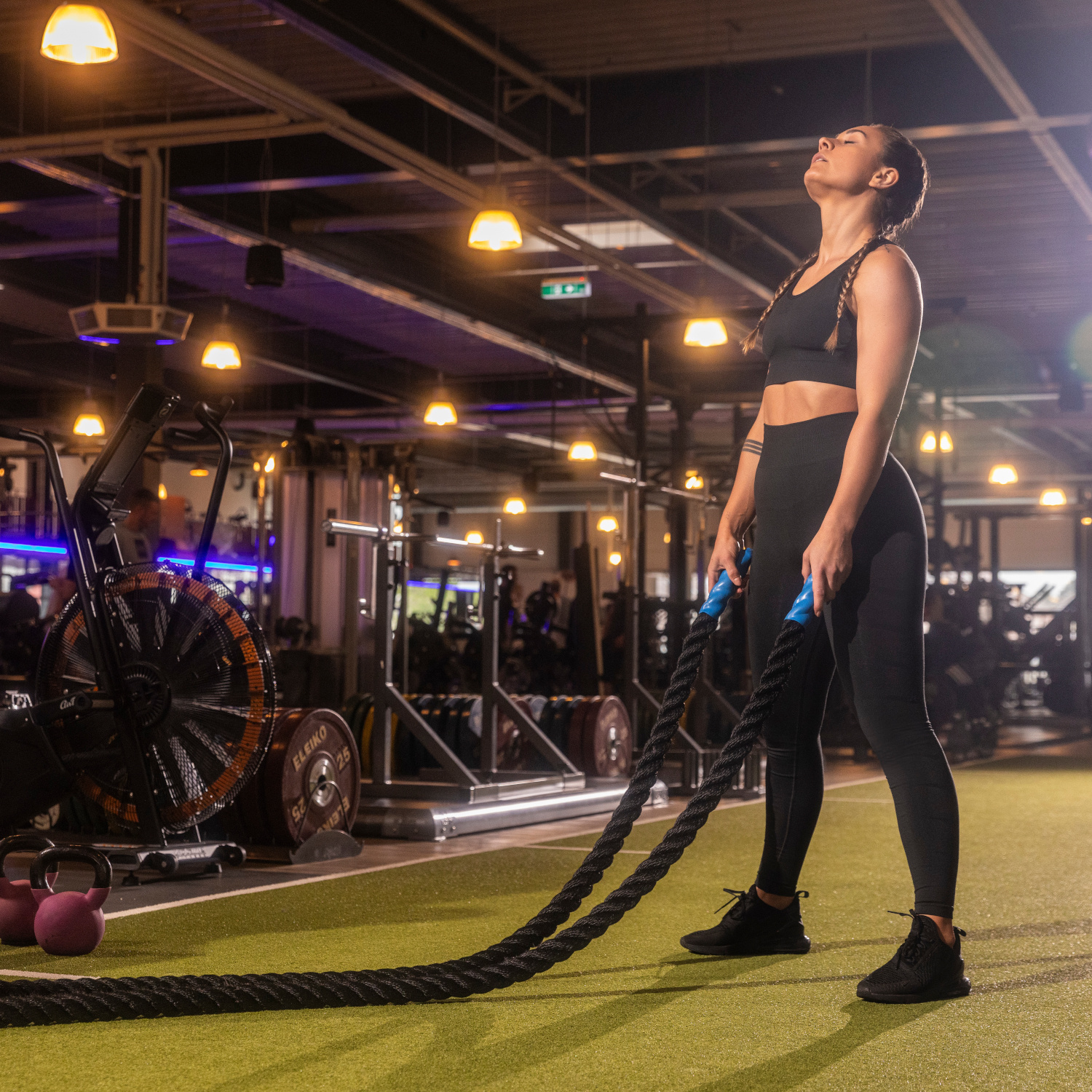 Eine Frau in Sportkleidung hält Trainingsseile und schaut nach oben, umgeben von Fitnessgeräten in einem gut beleuchteten Fitnessstudio.