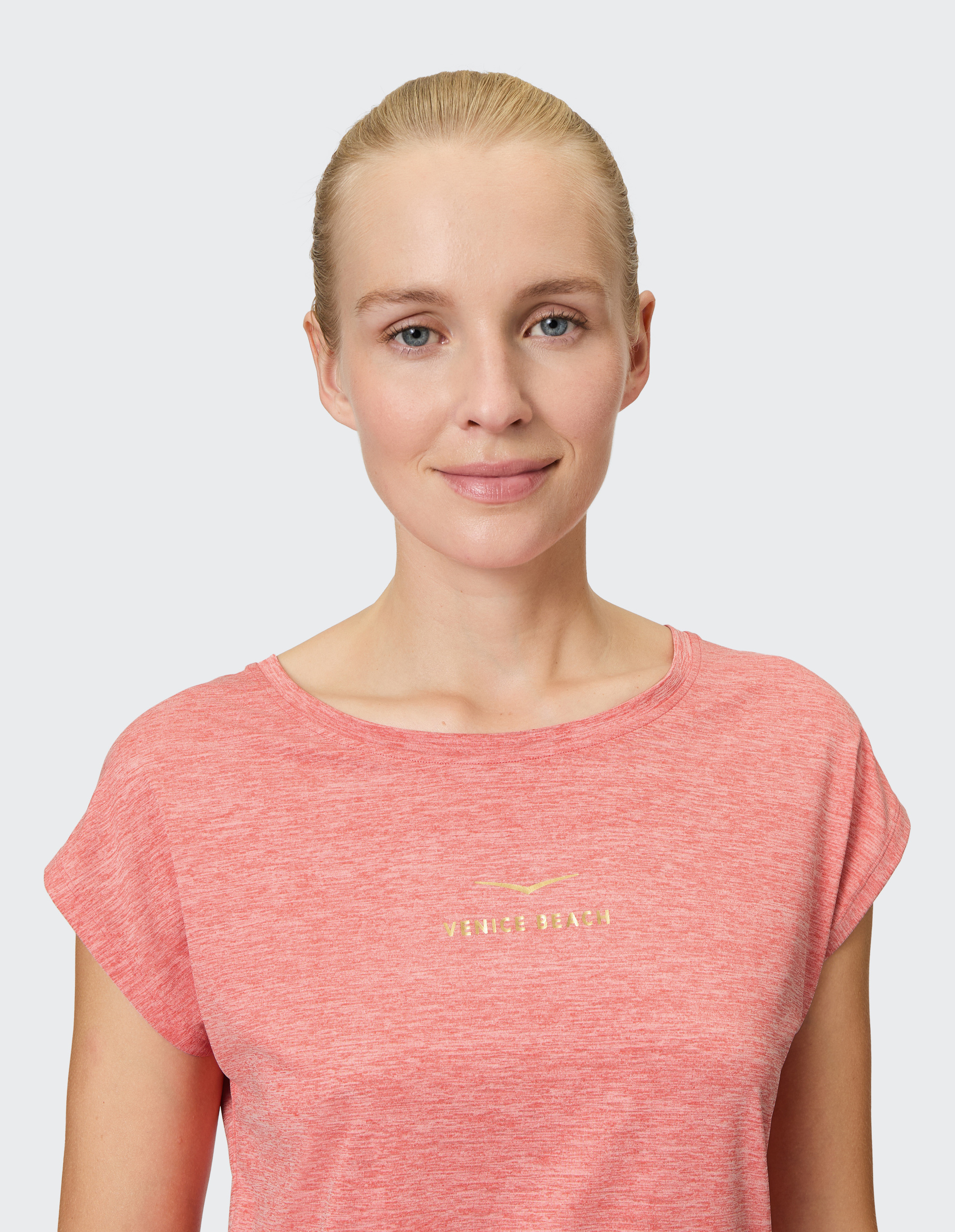 Frau mit blonden Haaren, lächelt sanft, trägt ein rotes T-Shirt mit der Aufschrift "Venice Beach" vor neutralem Hintergrund.