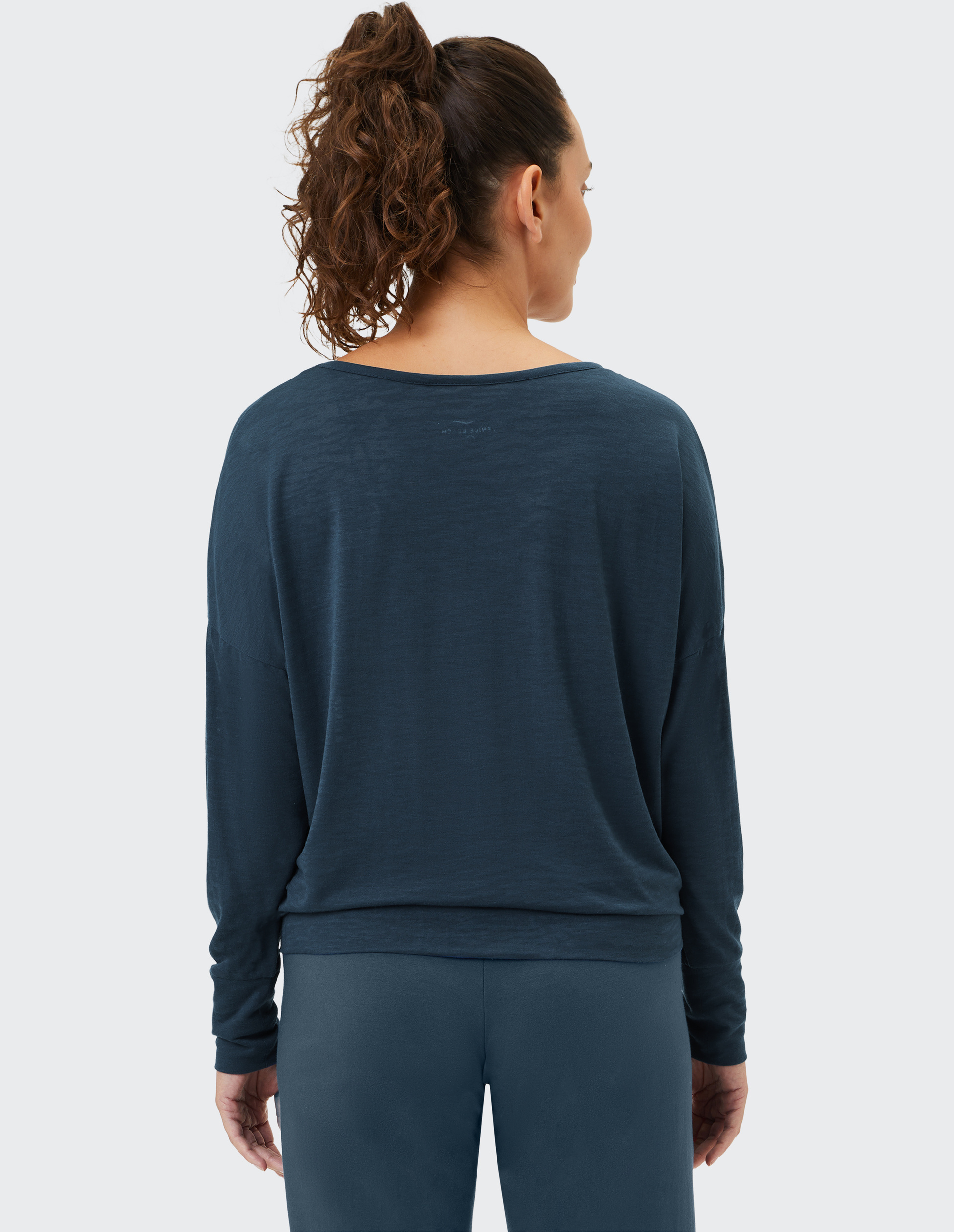 Eine Frau mit lockigem Pferdeschwanz trägt ein lockeres, dunkelblaues Langarmshirt und passende Leggings, von hinten betrachtet.