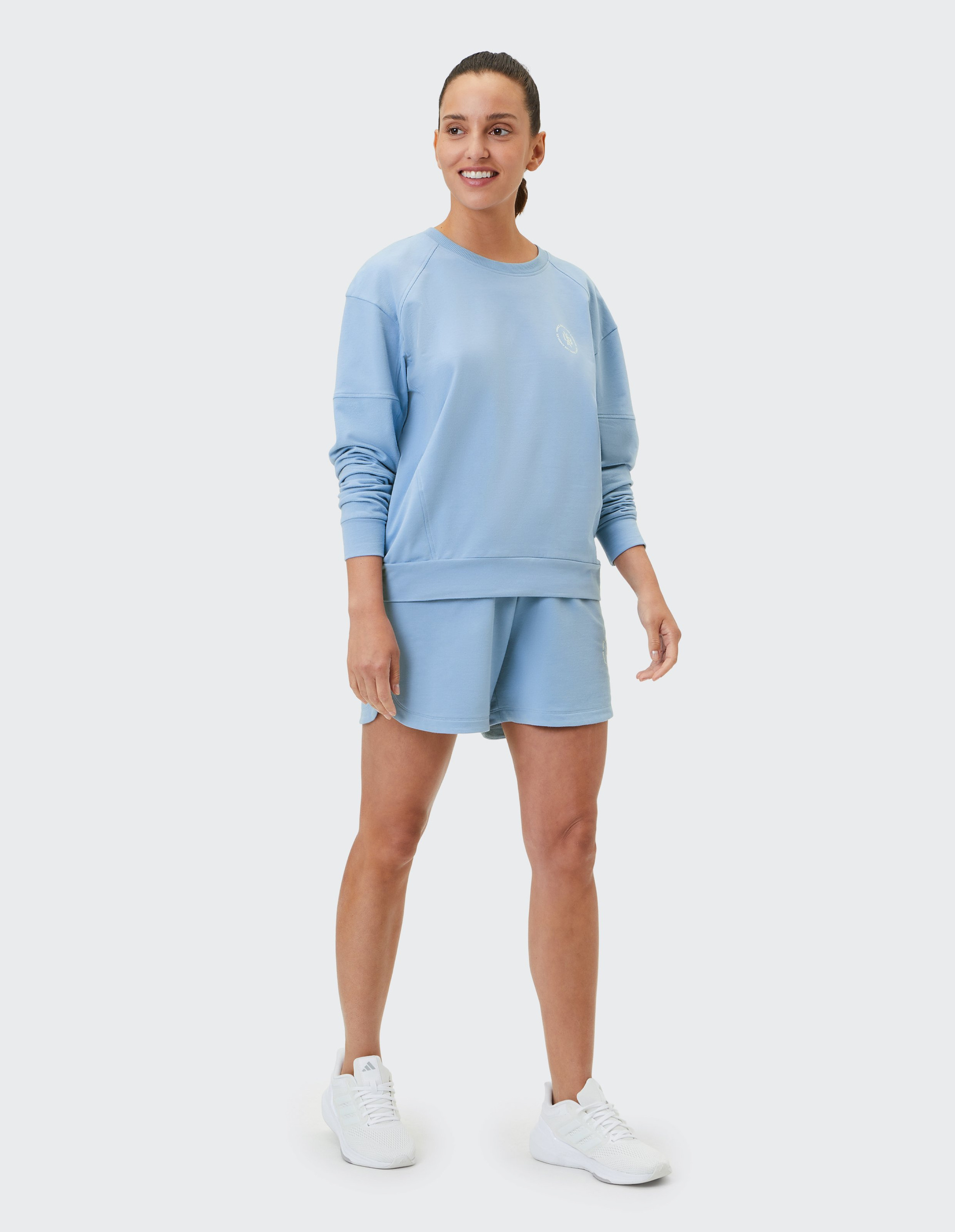Person in hellblauem Sweatshirt und Shorts steht lächelnd, trägt weiße Sneakers, hat braune Haare im Pferdeschwanz, auf neutralem Hintergrund.