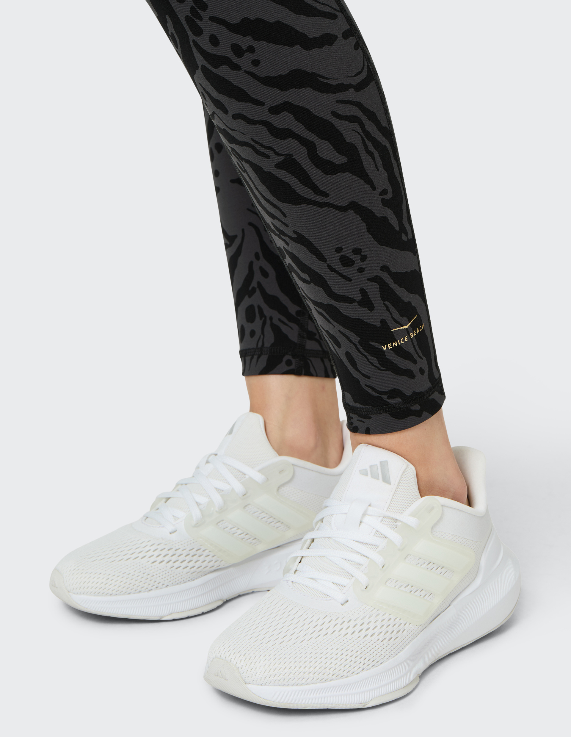 Weiße Sportschuhe mit Netzoberfläche und stabiler Sohle tragen, kombiniert mit schwarzen Leggings mit abstraktem Muster.