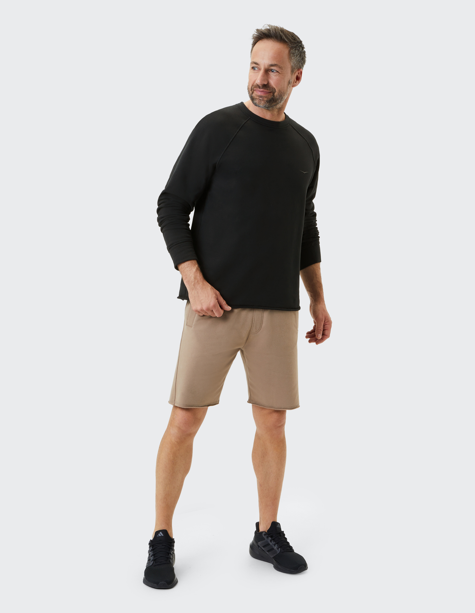 Ein Mann in einem schwarzen Langarmshirt und beigefarbenen Shorts steht lächelnd, blickt zur Seite, vor einem neutralen Hintergrund.