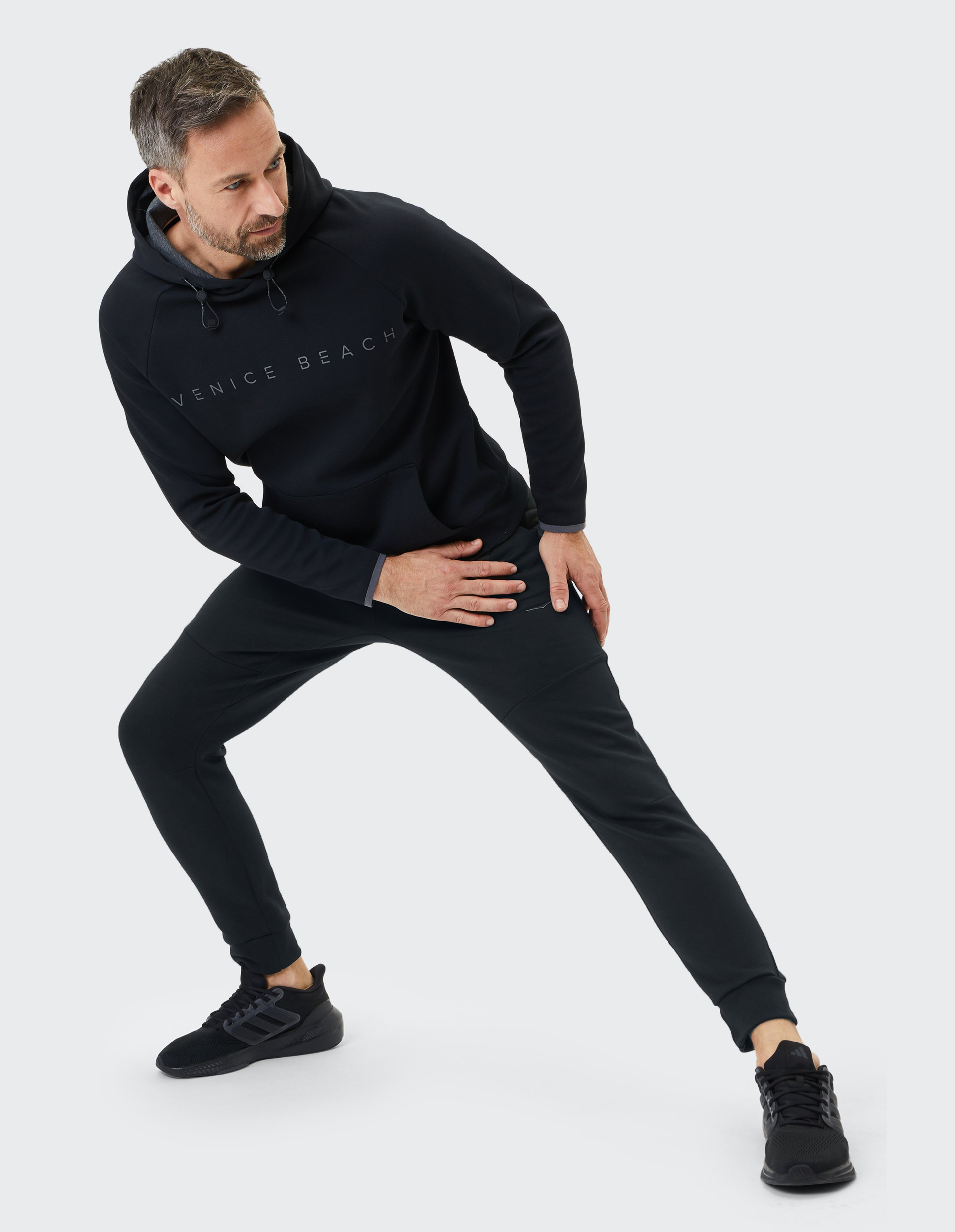 Ein Mann macht einen Ausfallschritt, trägt einen schwarzen Venice Beach Hoodie und Jogginghosen mit schwarzen Sneakern vor grauem Hintergrund.