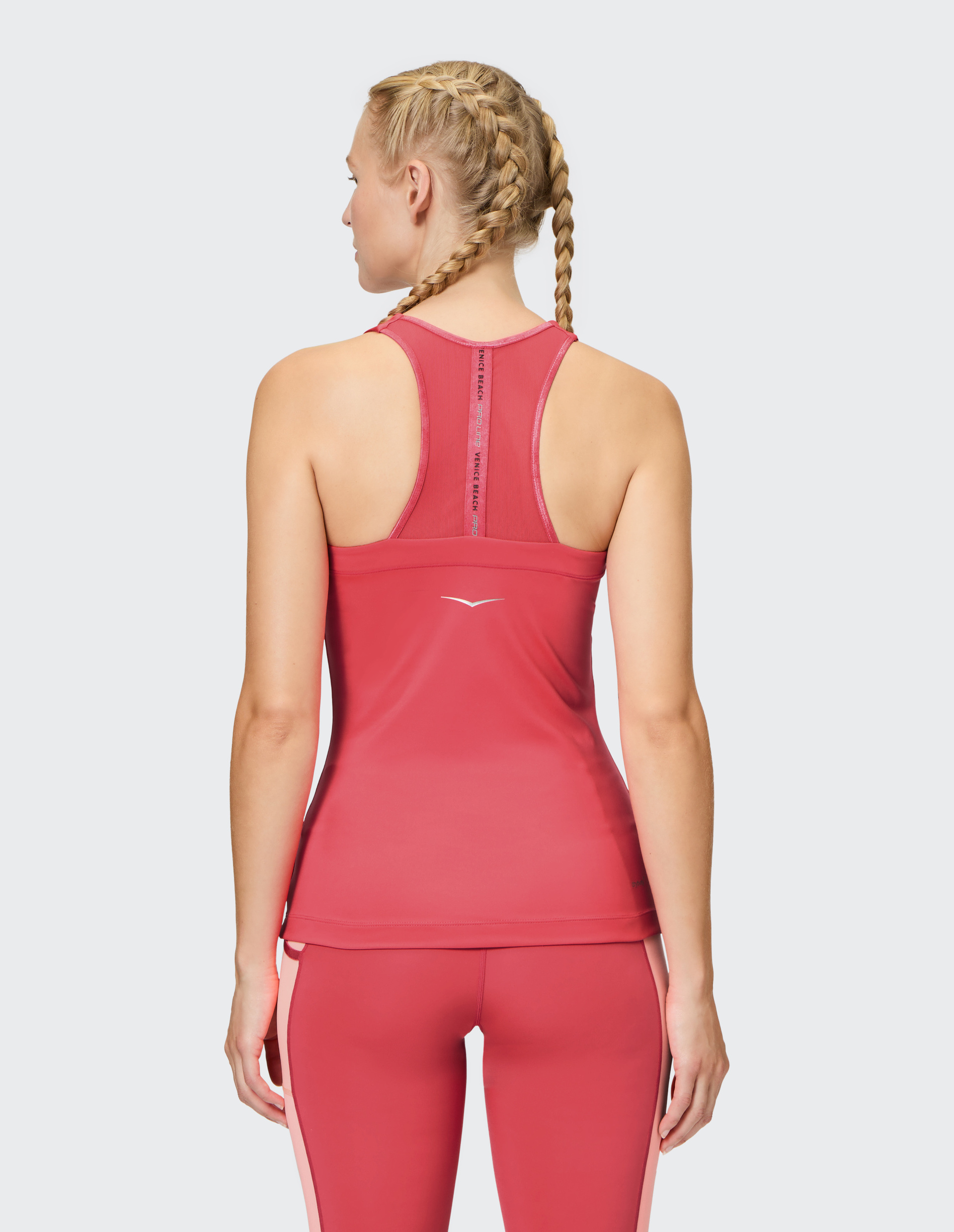 Frau von hinten in pinkem Sporttop und Leggings mit geflochtenem Haar; weißer Logo auf dem Oberteil, weißer Hintergrund.