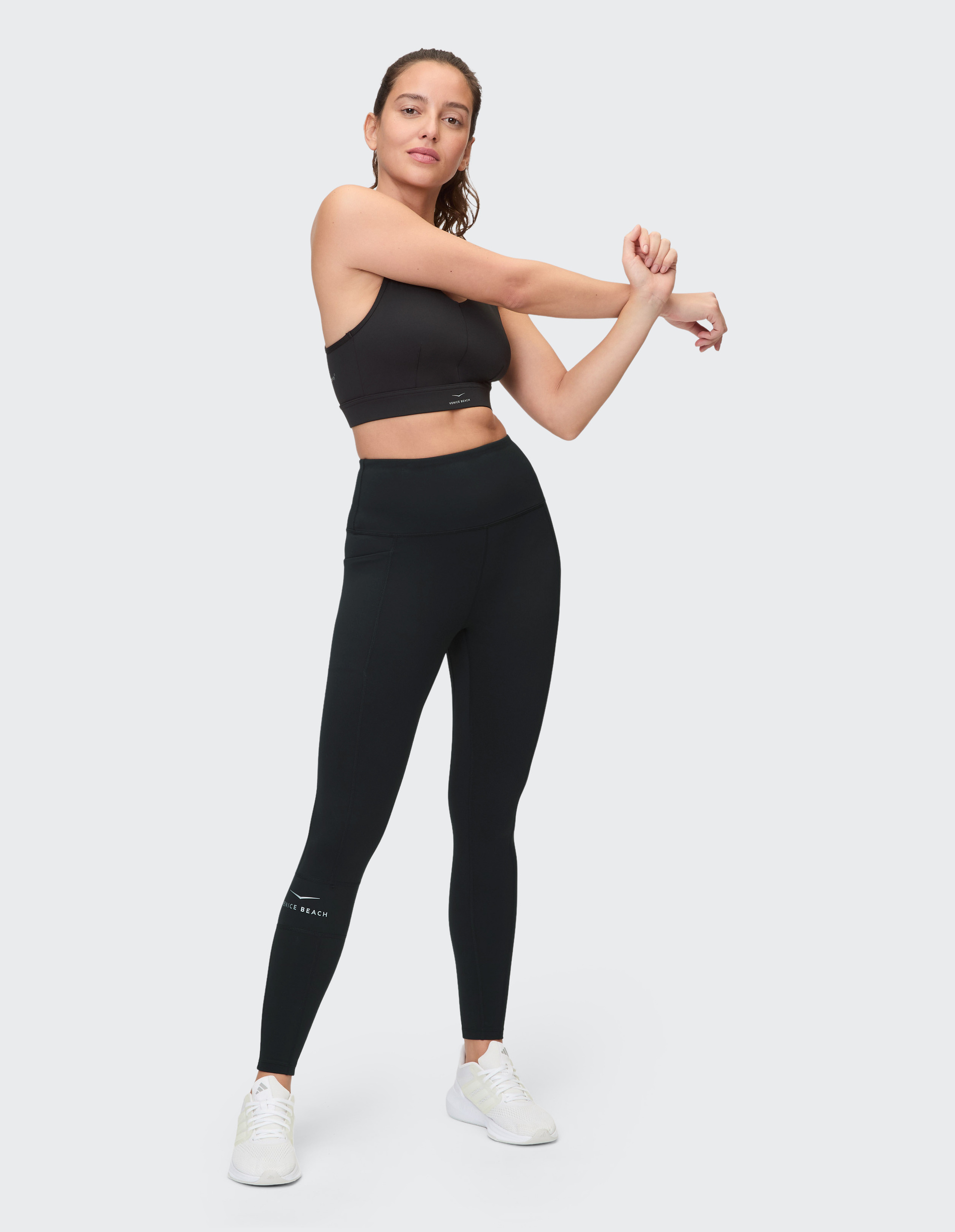 Frau in schwarzer Sportbekleidung mit Sport-BH und Leggings steht frontal, trägt weiße Turnschuhe und hat das Haar zurückgebunden.