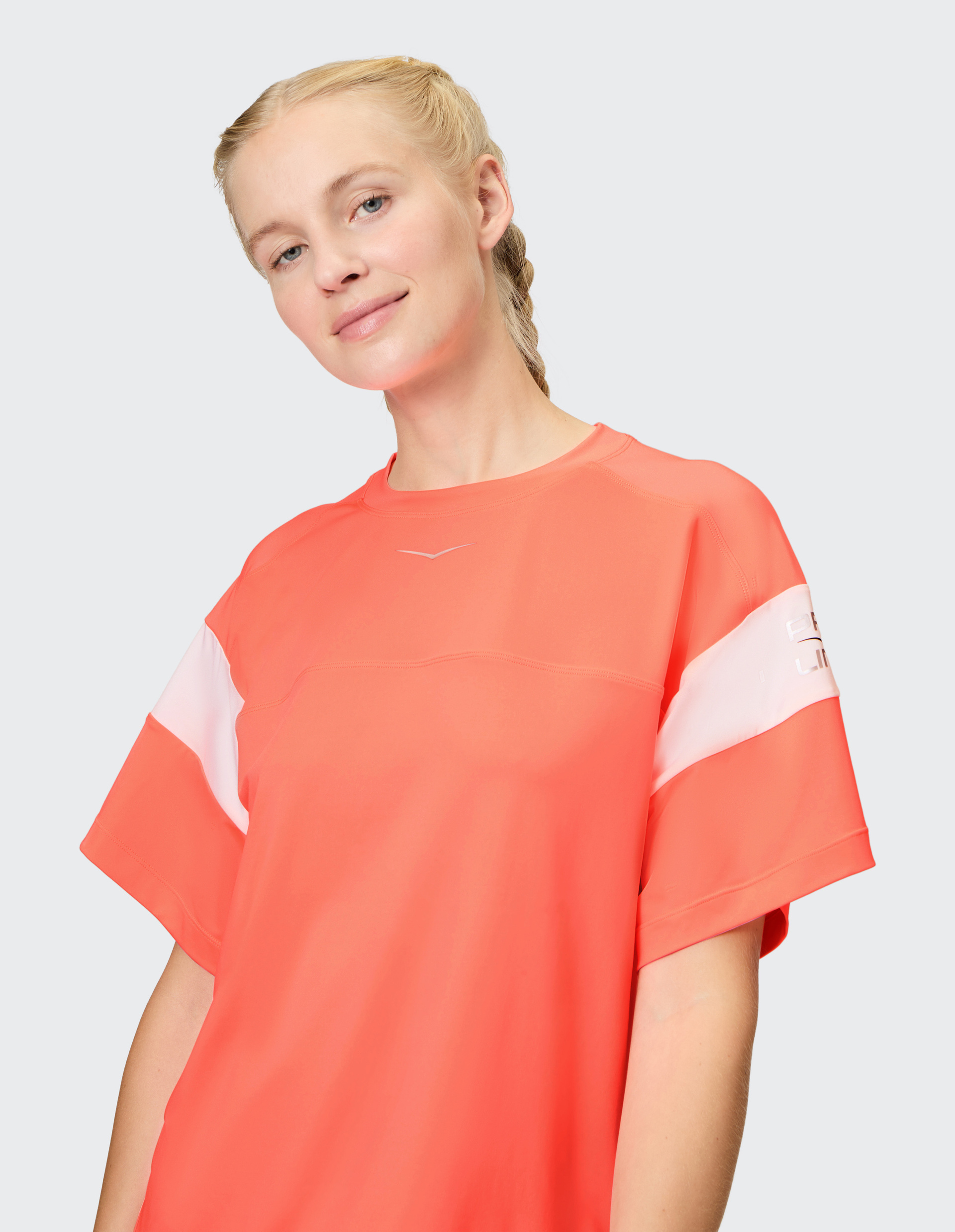 Frau mit blonden Zöpfen trägt ein leuchtend rotes T-Shirt mit weißen Streifen, lächelt leicht in Kamera auf grauem Hintergrund.