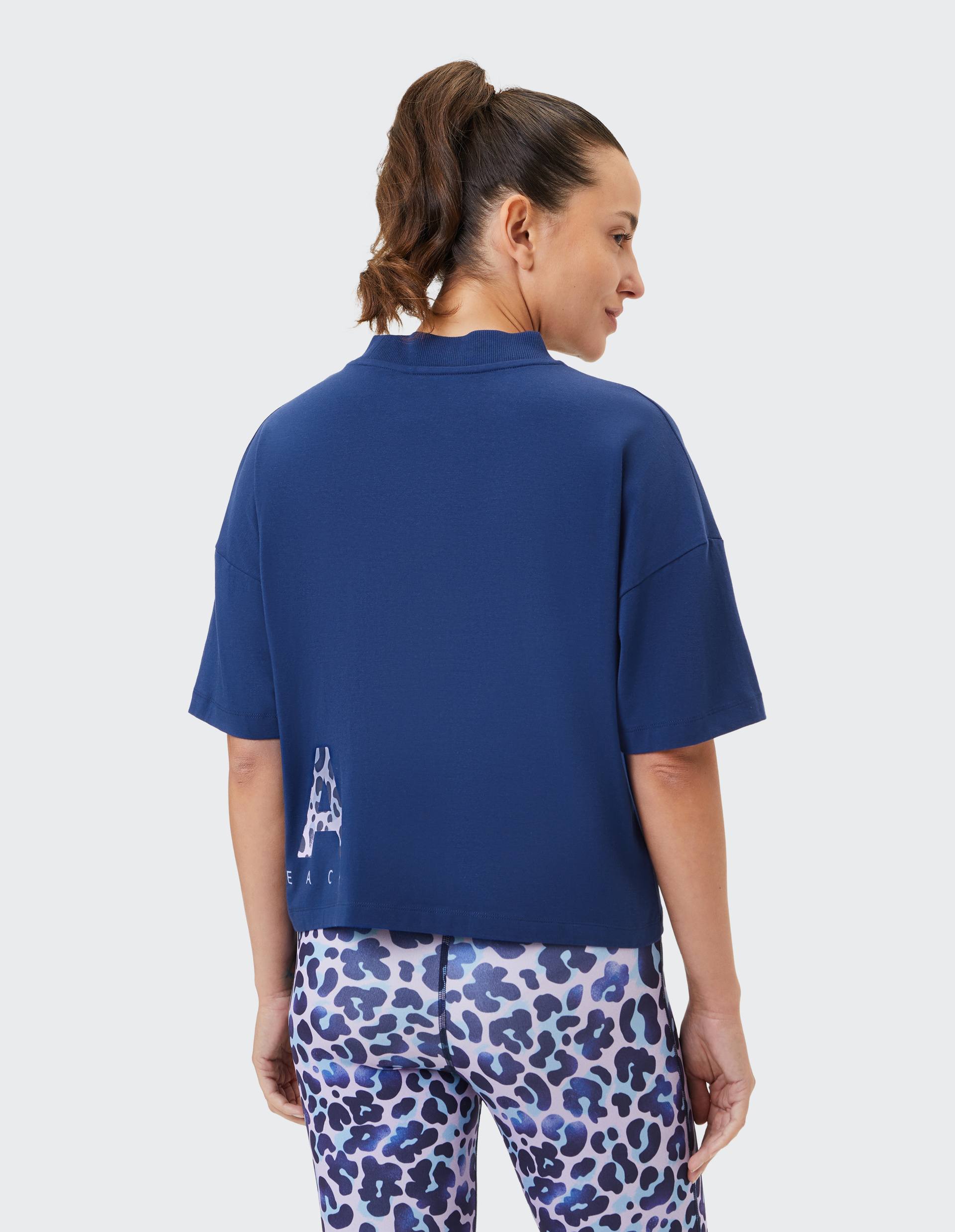 Frau mit Pferdeschwanz trägt blaues Shirt und bunte Leggings mit Leopardenmuster, steht von hinten vor hellem Hintergrund.