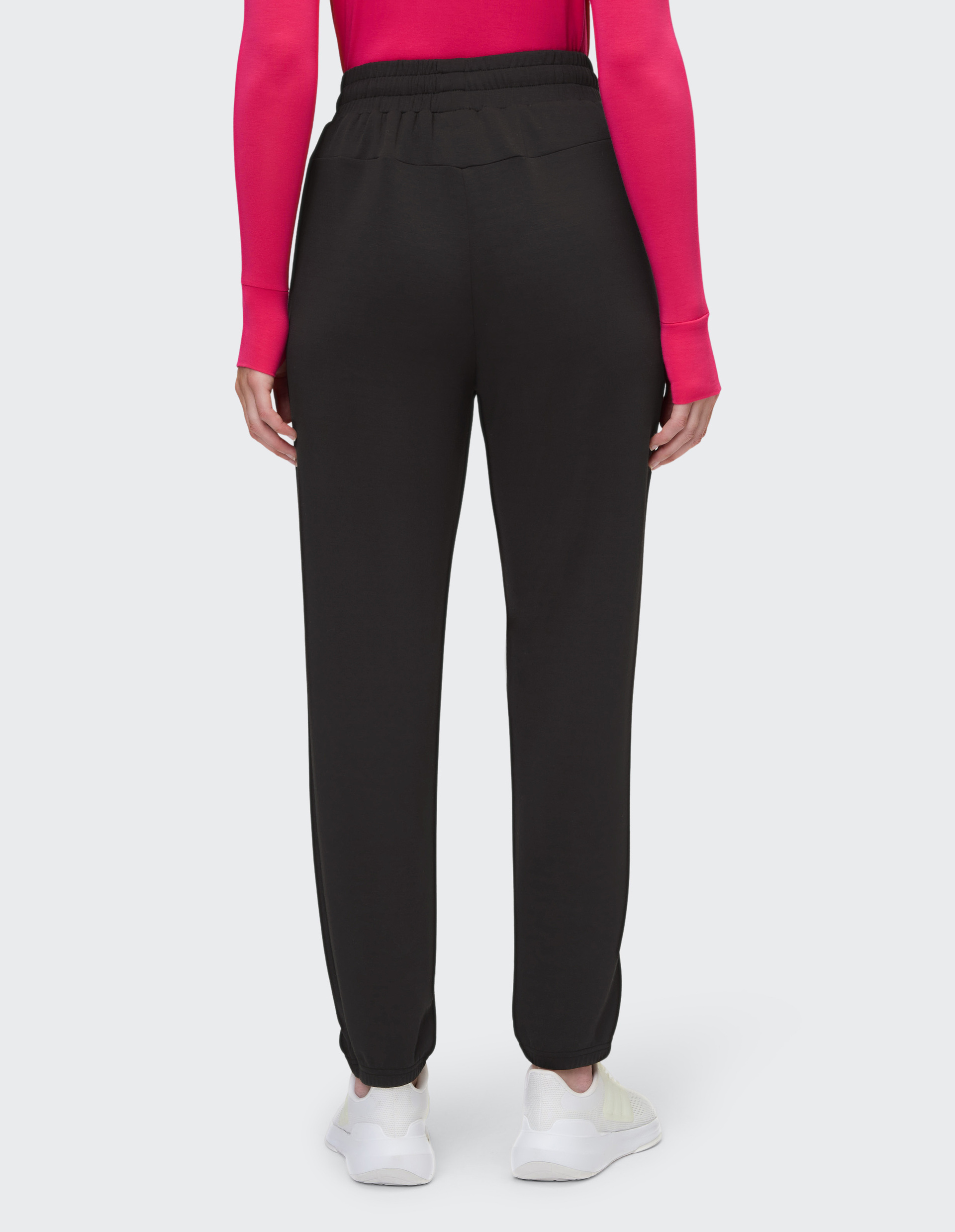 Person von hinten in schwarzer Hose und pinkem Langarmshirt, steht auf hellgrauem Hintergrund, trägt weiße Turnschuhe.