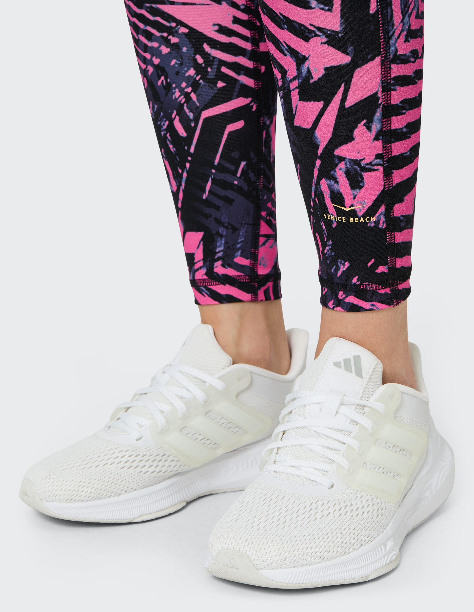 Weiße Turnschuhe auf grauem Boden, getragen zu farbenfrohen Leggings in Pink- und Schwarztönen mit geometrischem Muster.
