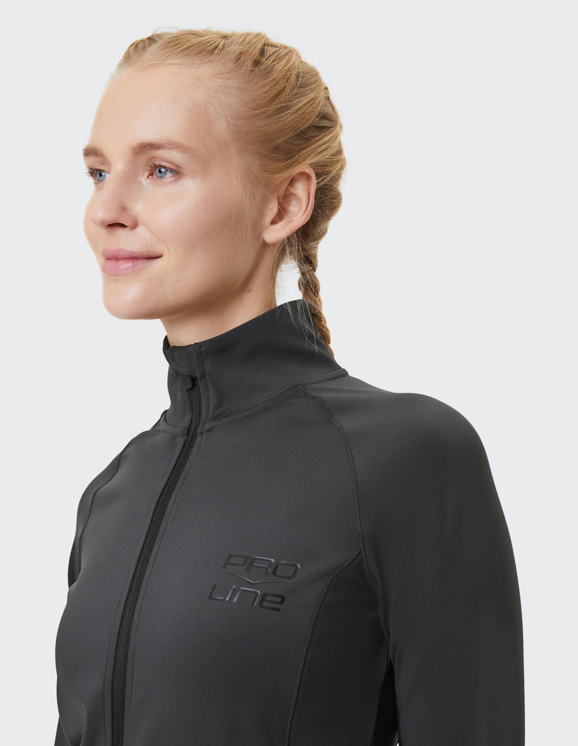 Eine lächelnde Frau mit geflochtenem Haar trägt eine schwarze Sportjacke mit Pro Line-Logo vor einem hellen Hintergrund.