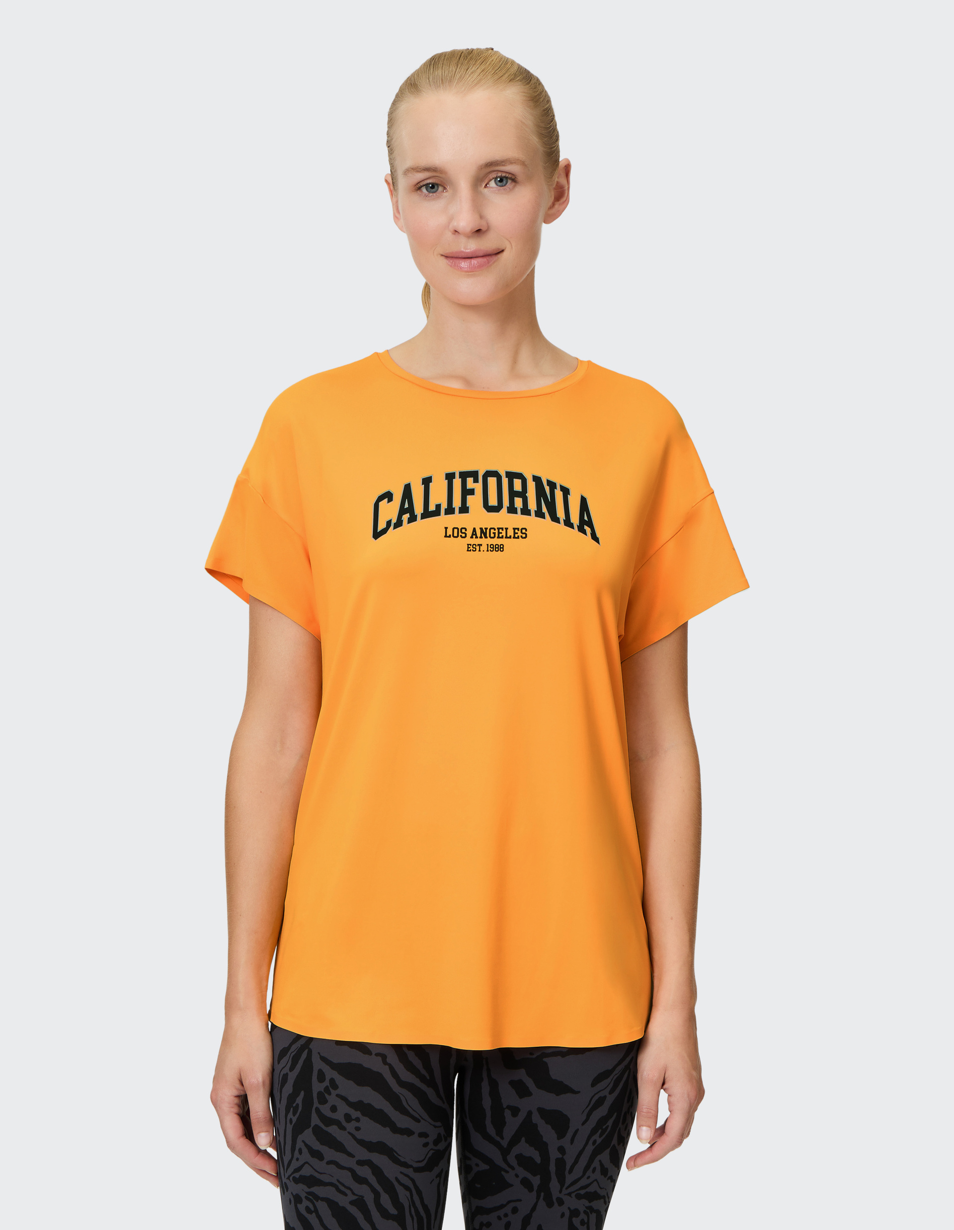 Frau trägt ein orangefarbenes T-Shirt mit Aufdruck "California Los Angeles" und dunkle gemusterte Hose vor hellem Hintergrund.