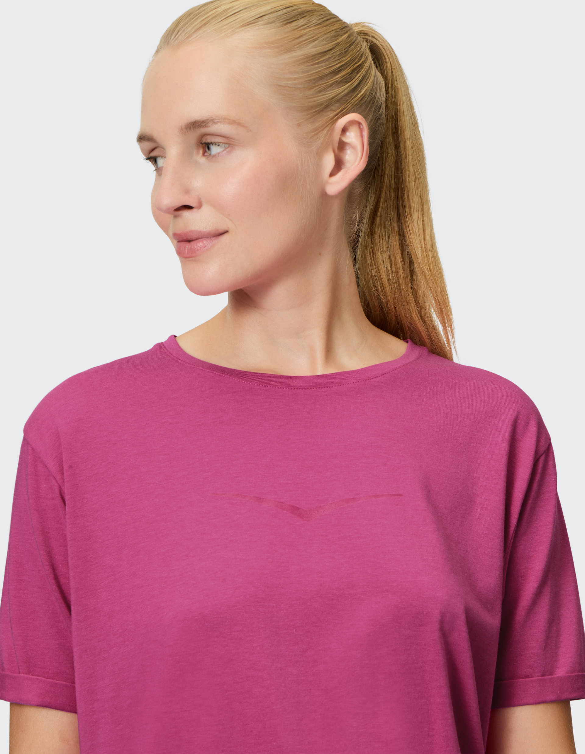 Eine Frau mit blonden Haaren im Pferdeschwanz trägt ein rosa T-Shirt und blickt lächelnd zur Seite vor grauem Hintergrund.