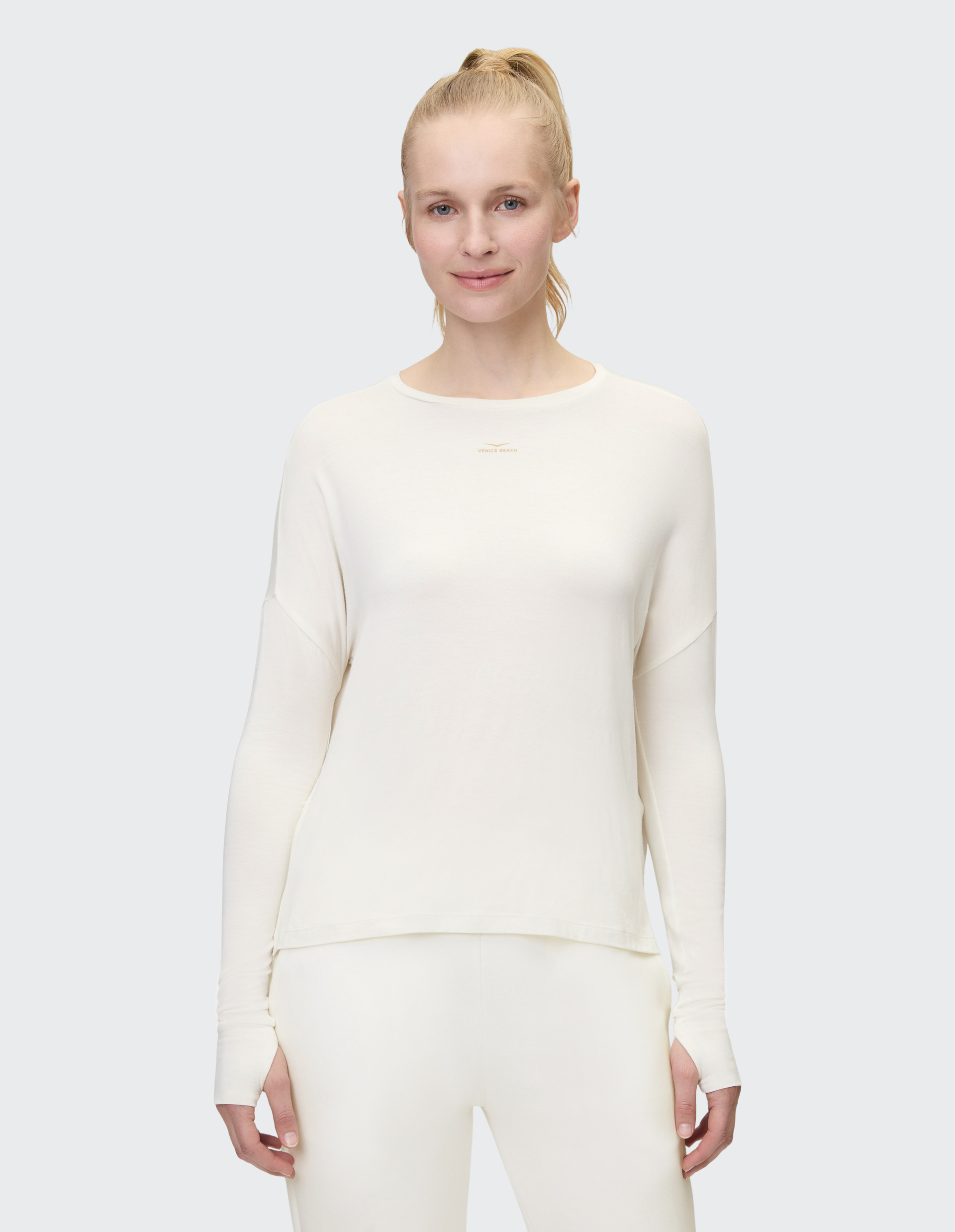 Eine Person mit blonden Haaren trägt ein schlichtes, weißes Langarm-Oberteil und passende weiße Hosen, vor grauem Hintergrund.