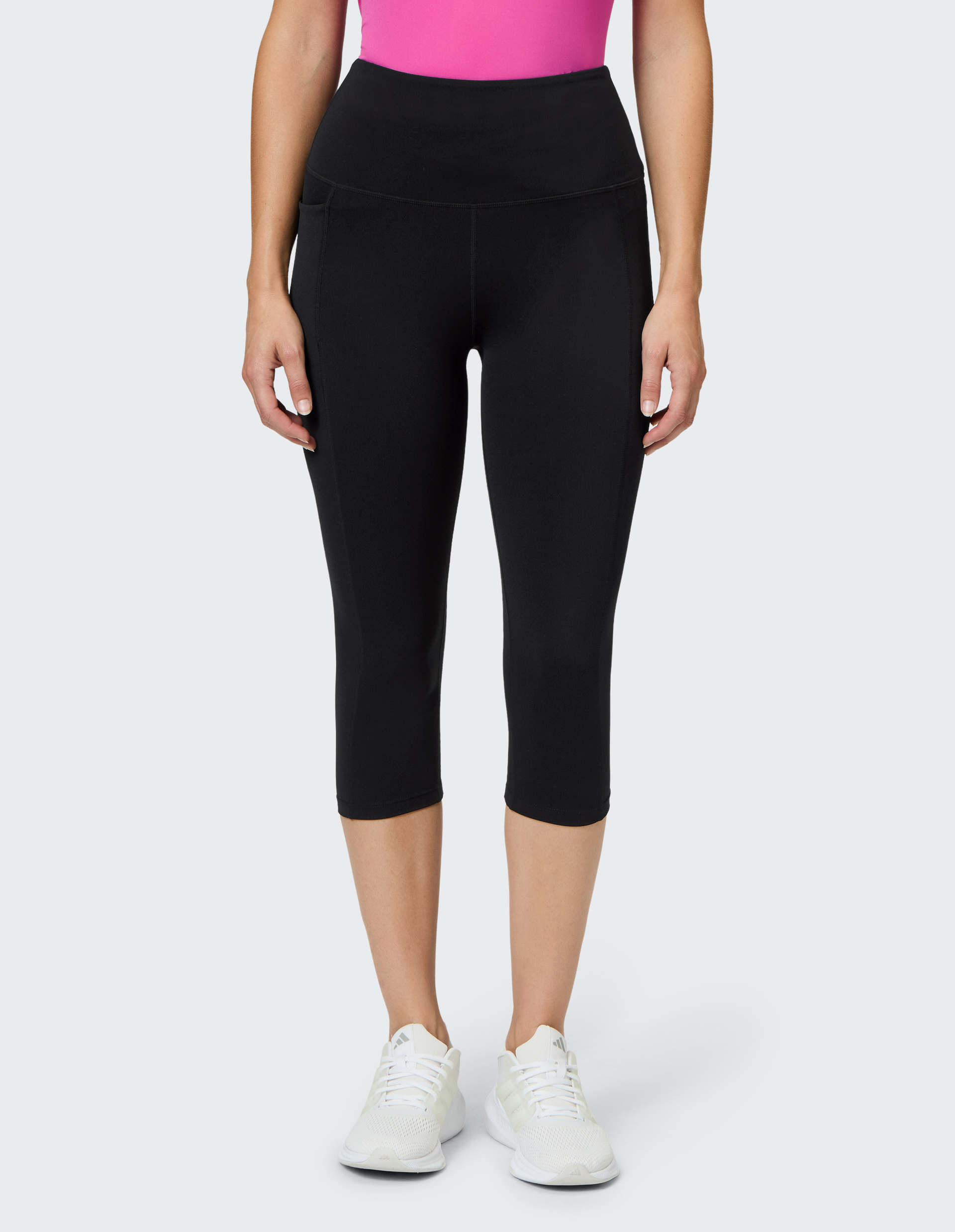 Eine Person trägt ein pinkes Oberteil, schwarze Capri-Leggings und weiße Sportschuhe auf grauem Hintergrund.