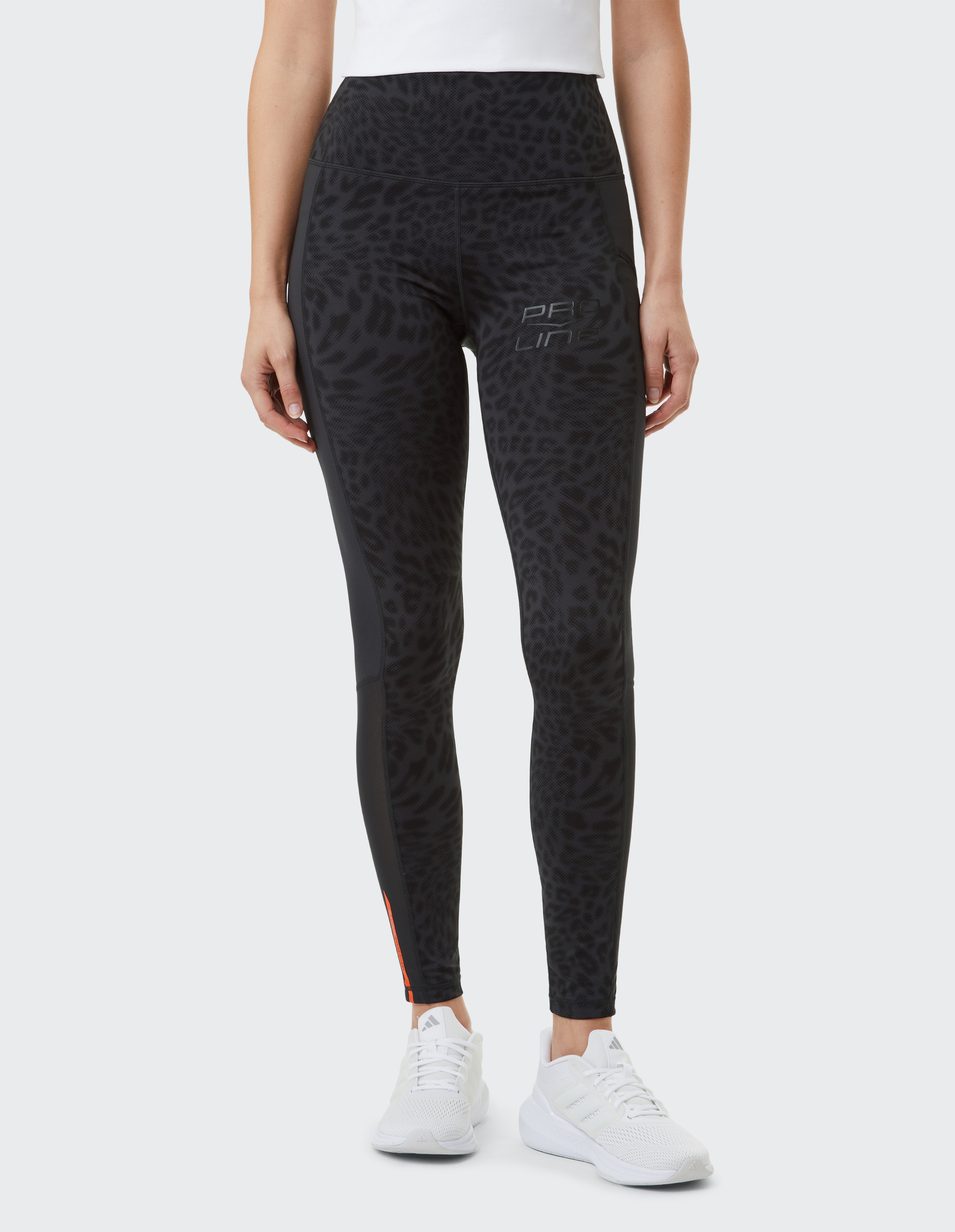 Frau in schwarzer Leggings mit Leopardenmuster und weißem Shirt, trägt weiße Sportschuhe, steht vor neutralem Hintergrund.