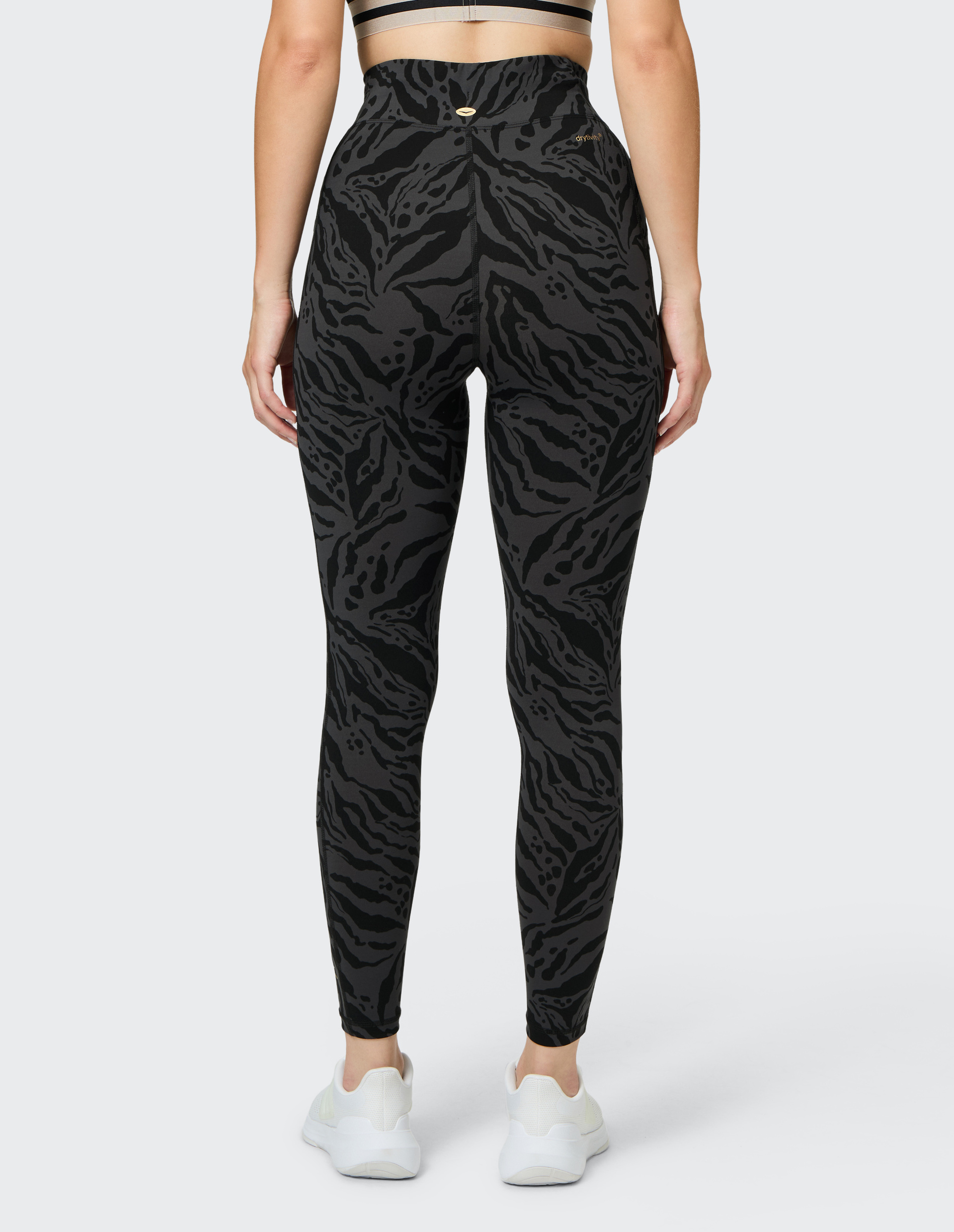 Rückansicht einer Frau in schwarz gemusterten Leggings mit hohem Bund und Sneakers, die einen hellen Sport-BH trägt.