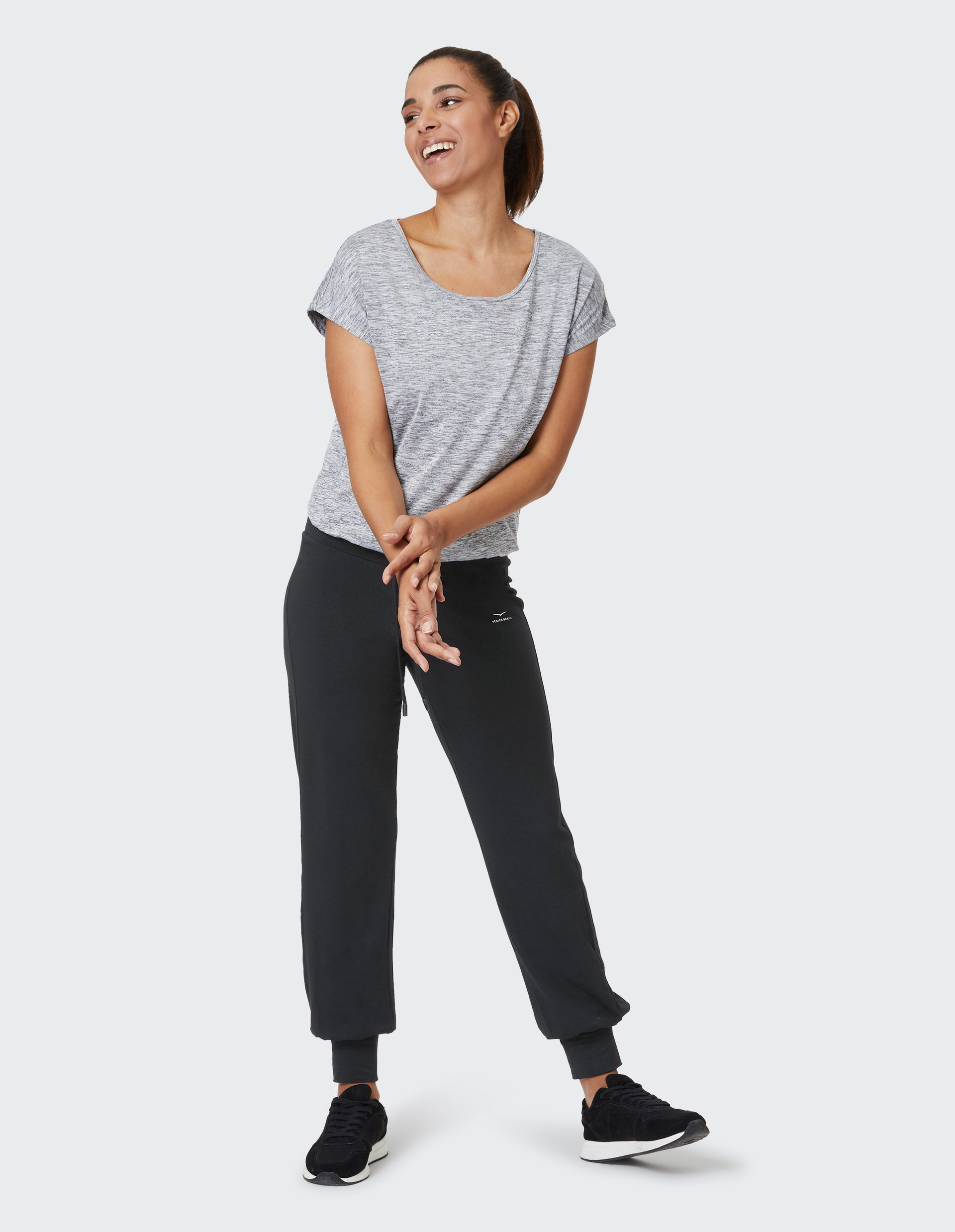 Eine Frau in sportlicher Kleidung, grauem T-Shirt und schwarzer Jogginghose, lächelnd und locker stehend vor weißem Hintergrund.