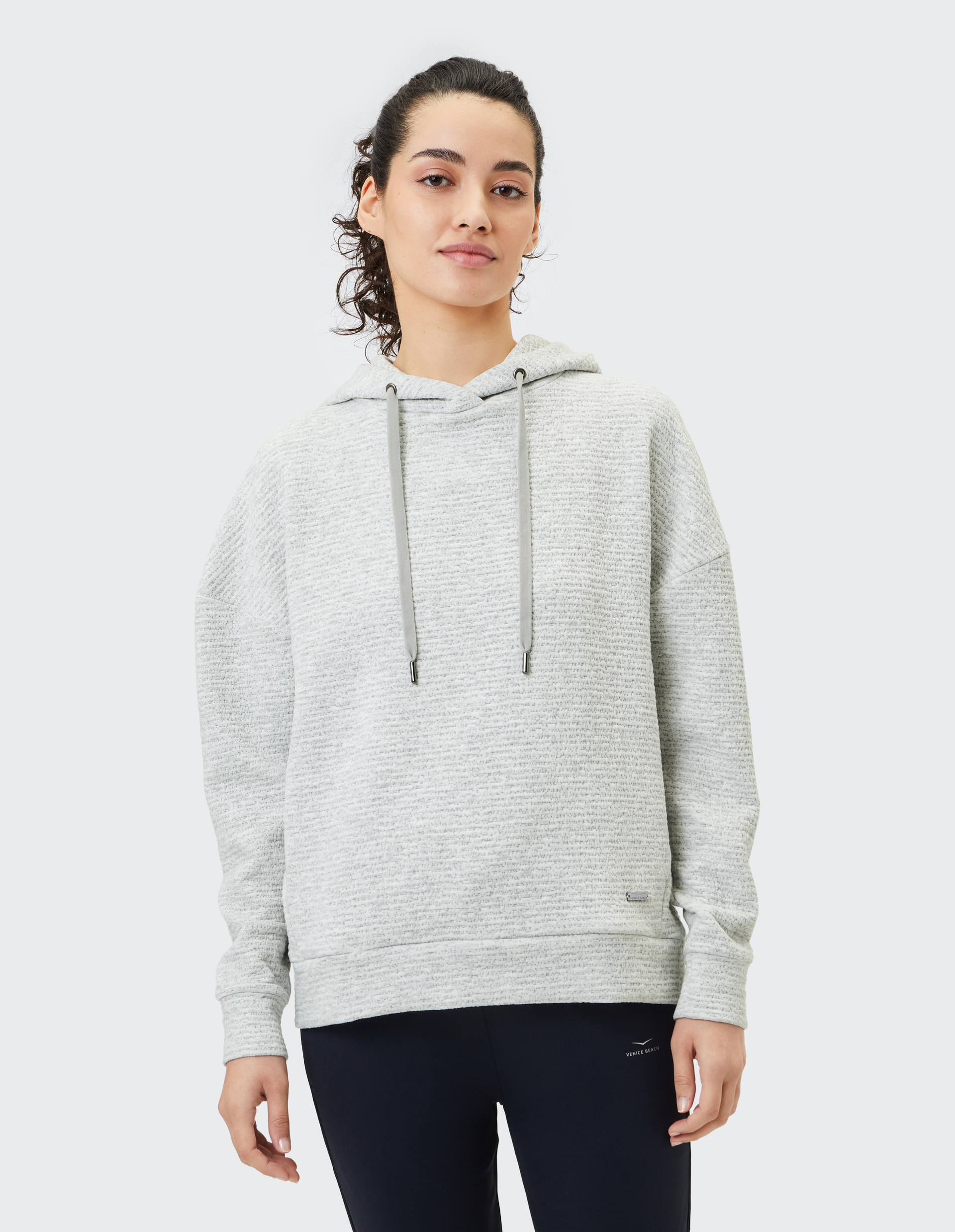 Frau in grauem Kapuzenpullover mit Kordeln und schwarzer Hose vor hellem Hintergrund, schaut freundlich in die Kamera.