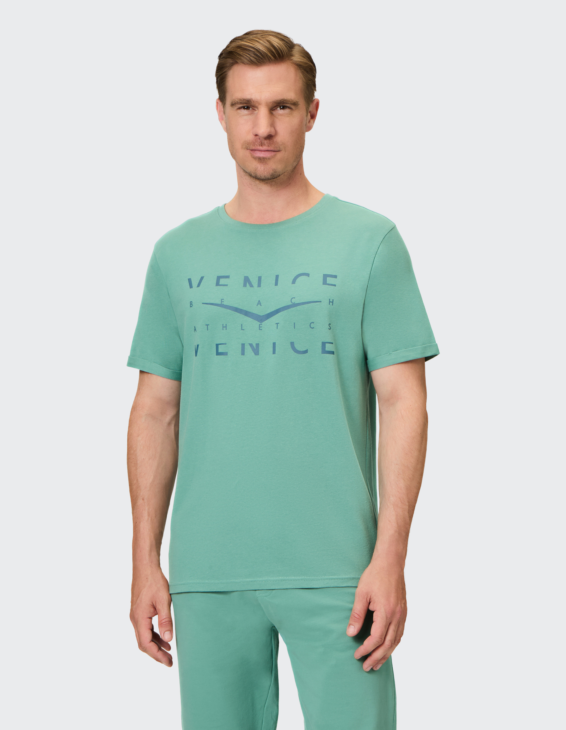 Mann in grünem T-Shirt und Hose mit VENICE Beach Athletics-Aufdruck, steht vor hellem Hintergrund, schaut freundlich zur Kamera.