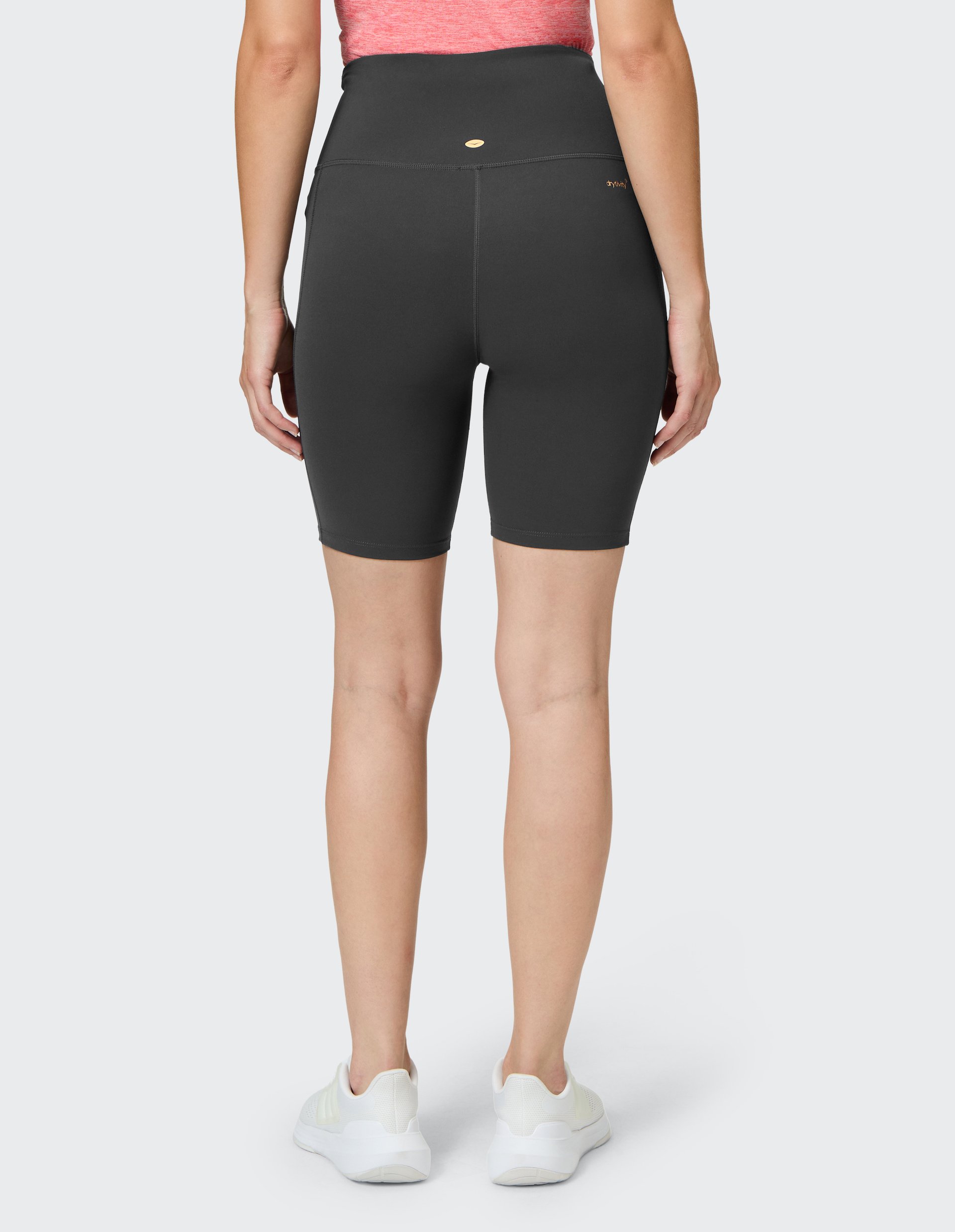 Frau in schwarzer, hochtaillierter Radlerhose, rotem Oberteil und weißen Turnschuhen, von hinten, steht vor grauem Hintergrund.