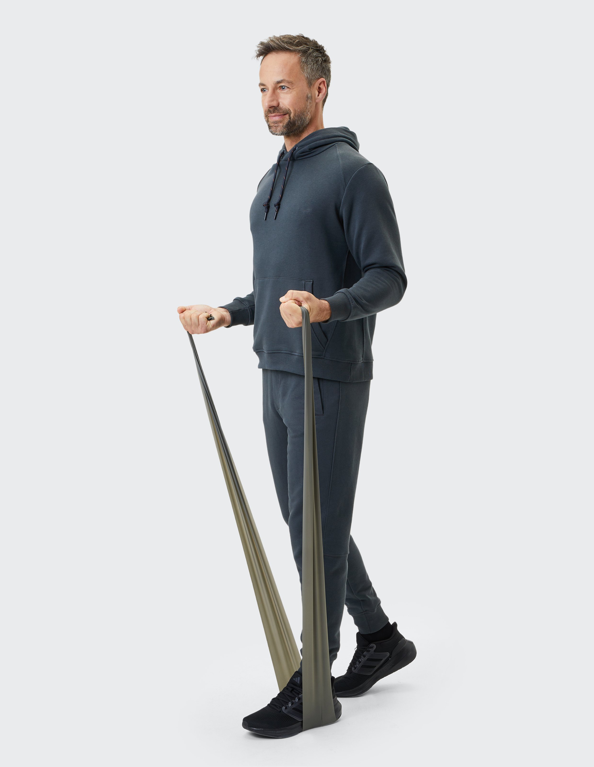 Ein Mann in dunkelgrauem Trainingsanzug trainiert mit einem Fitnessband, das unter seinen Füßen gespannt ist, vor hellem Hintergrund.