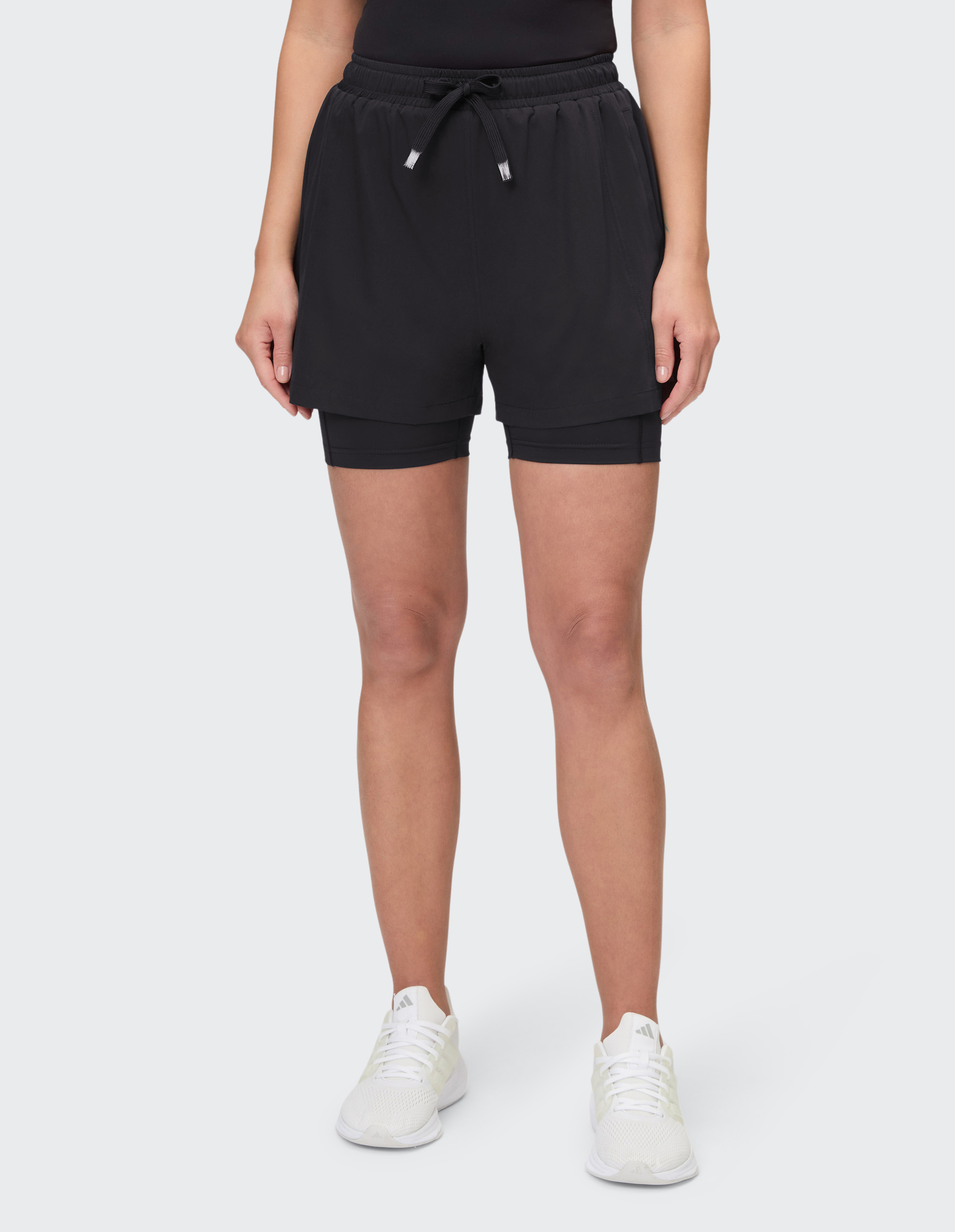 Frau trägt schwarze Shorts mit Kordelzug und weißen Sneakers auf hellem Hintergrund, oberer Körper nicht sichtbar.