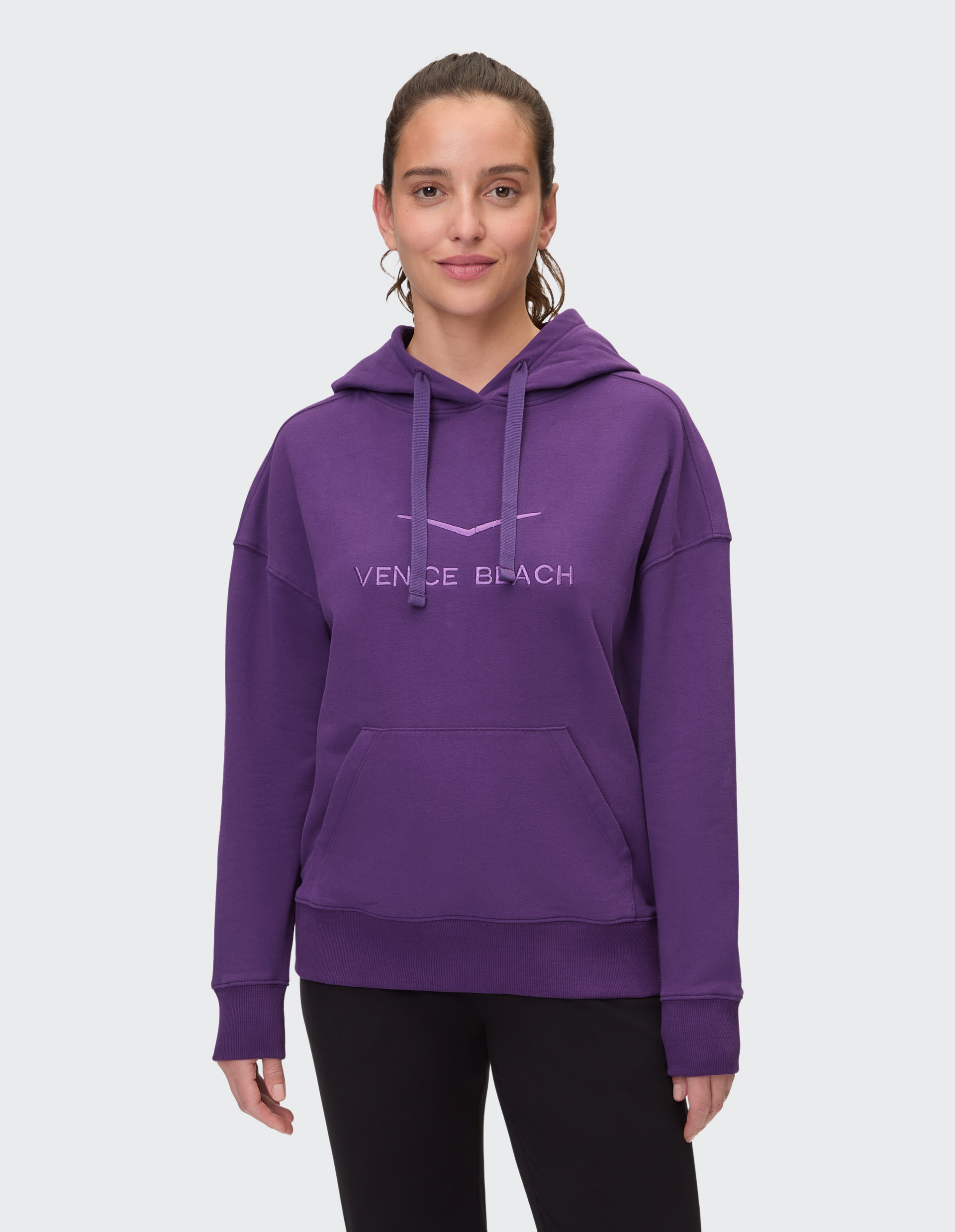 Frau in lila Kapuzenpullover mit Venice Beach-Schriftzug, lächelt, dunkle Hose, heller Hintergrund.