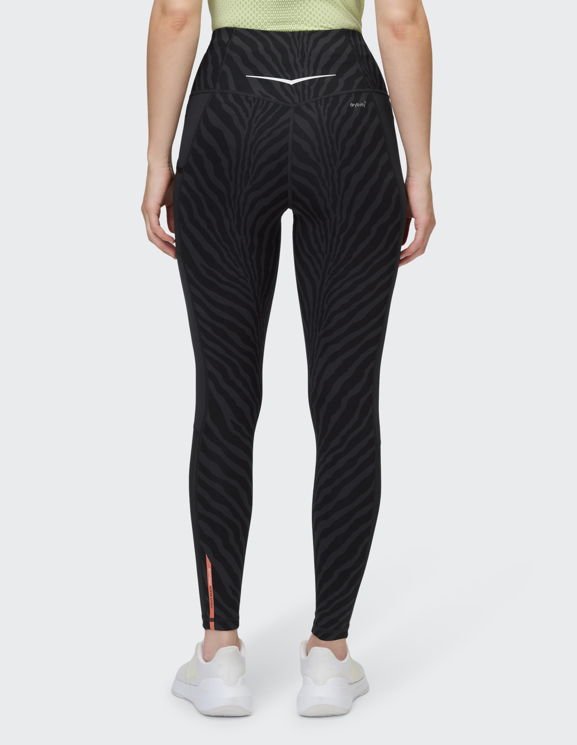 Frau in grüner Mesh-Oberteil, trägt schwarze Leggings mit Zebramuster und weißen Turnschuhen, von hinten gesehen.