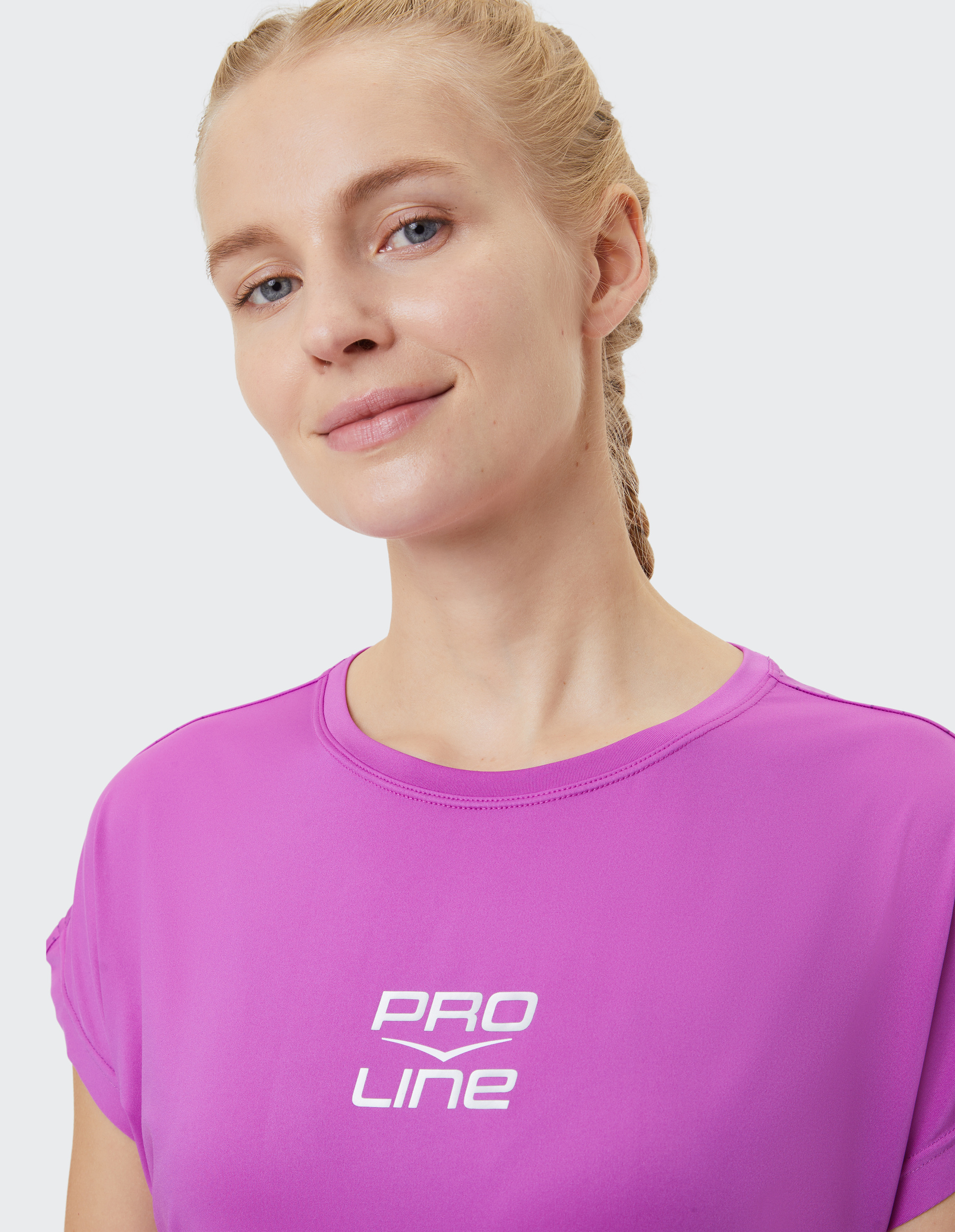 Frau mit blondem Zopf trägt ein lila Shirt mit der Aufschrift "Pro Line" und lächelt leicht in die Kamera vor neutralem Hintergrund.