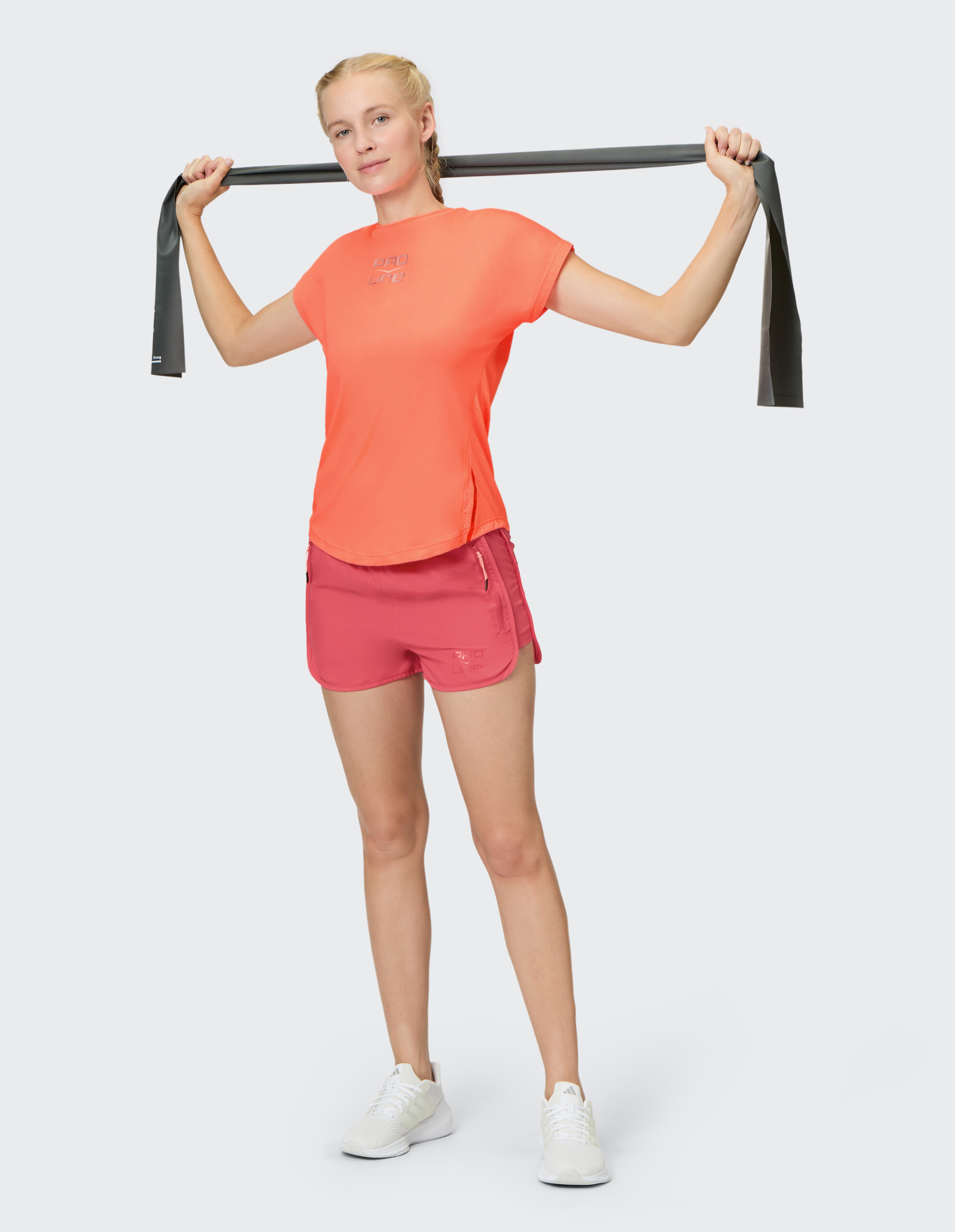 Frau in orangefarbenem Shirt und roten Shorts hält ein schwarzes Fitnessband über den Schultern, trägt weiße Sneakers.