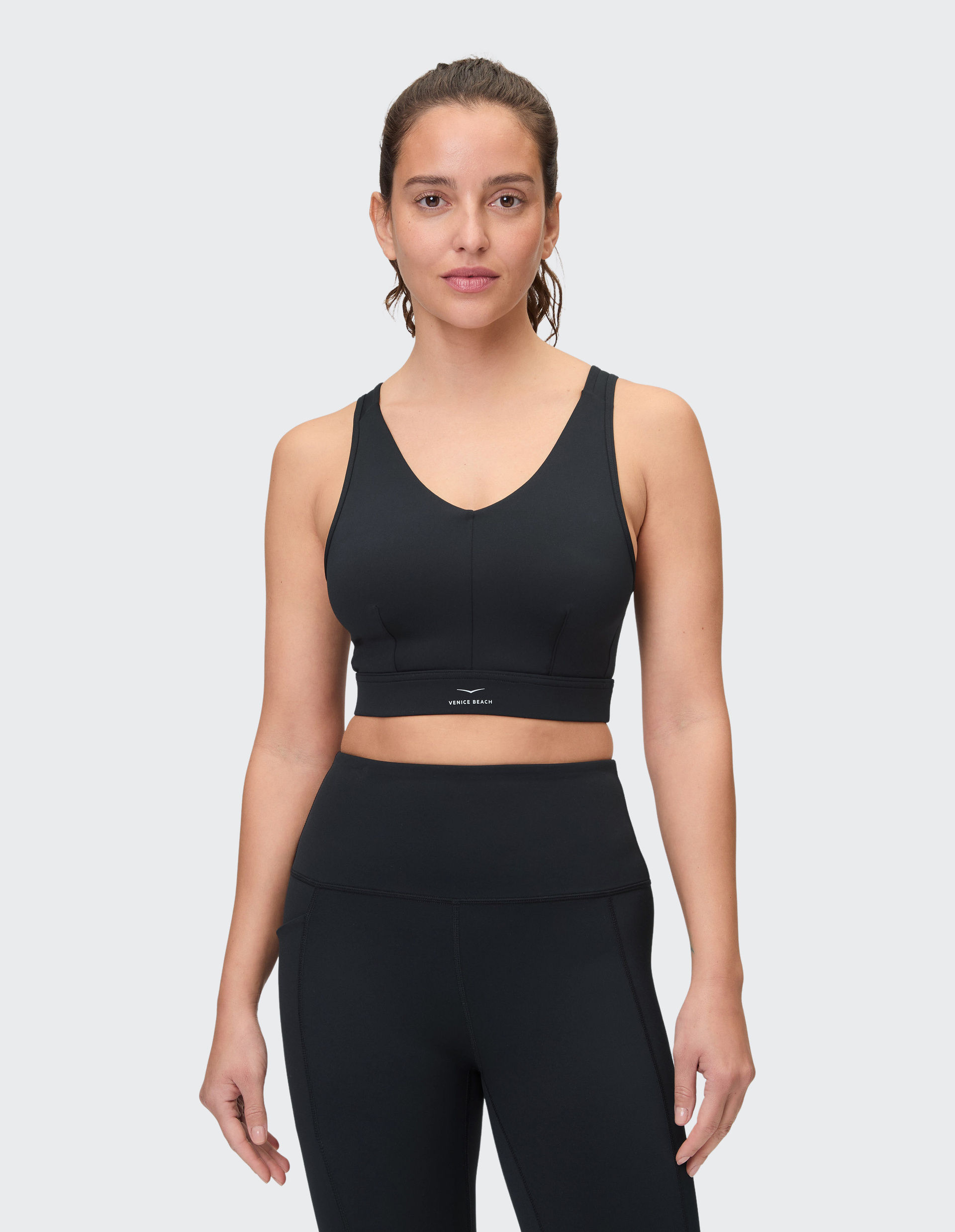 Eine Frau trägt ein schwarzes Sporttop und Leggings, steht frontal mit neutralem Gesichtsausdruck vor einem hellgrauen Hintergrund.