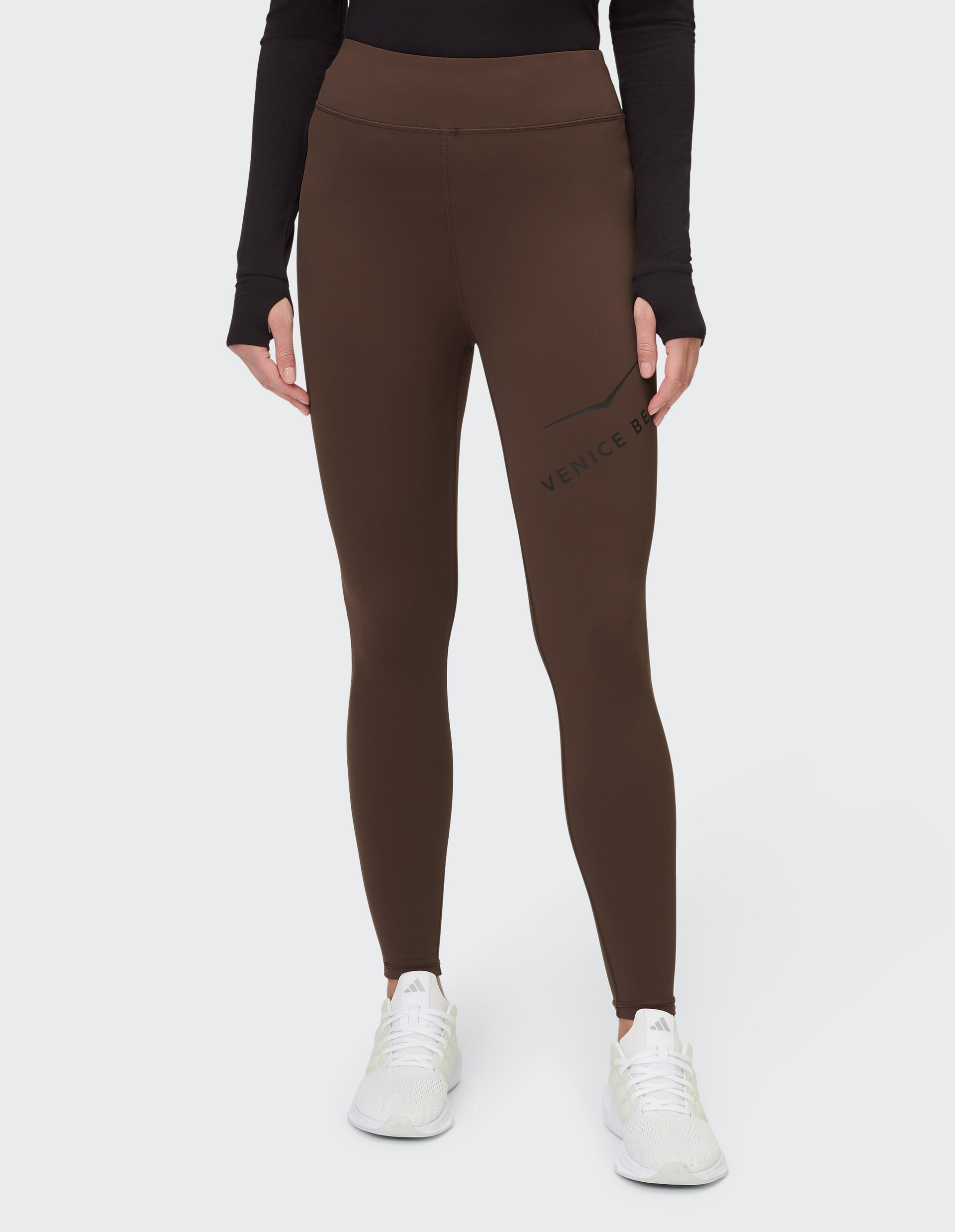 Person trägt eine braune Leggings mit "Venice Beach"-Logo, schwarzem Langarmshirt und weißen Sneakers, vor weißem Hintergrund.