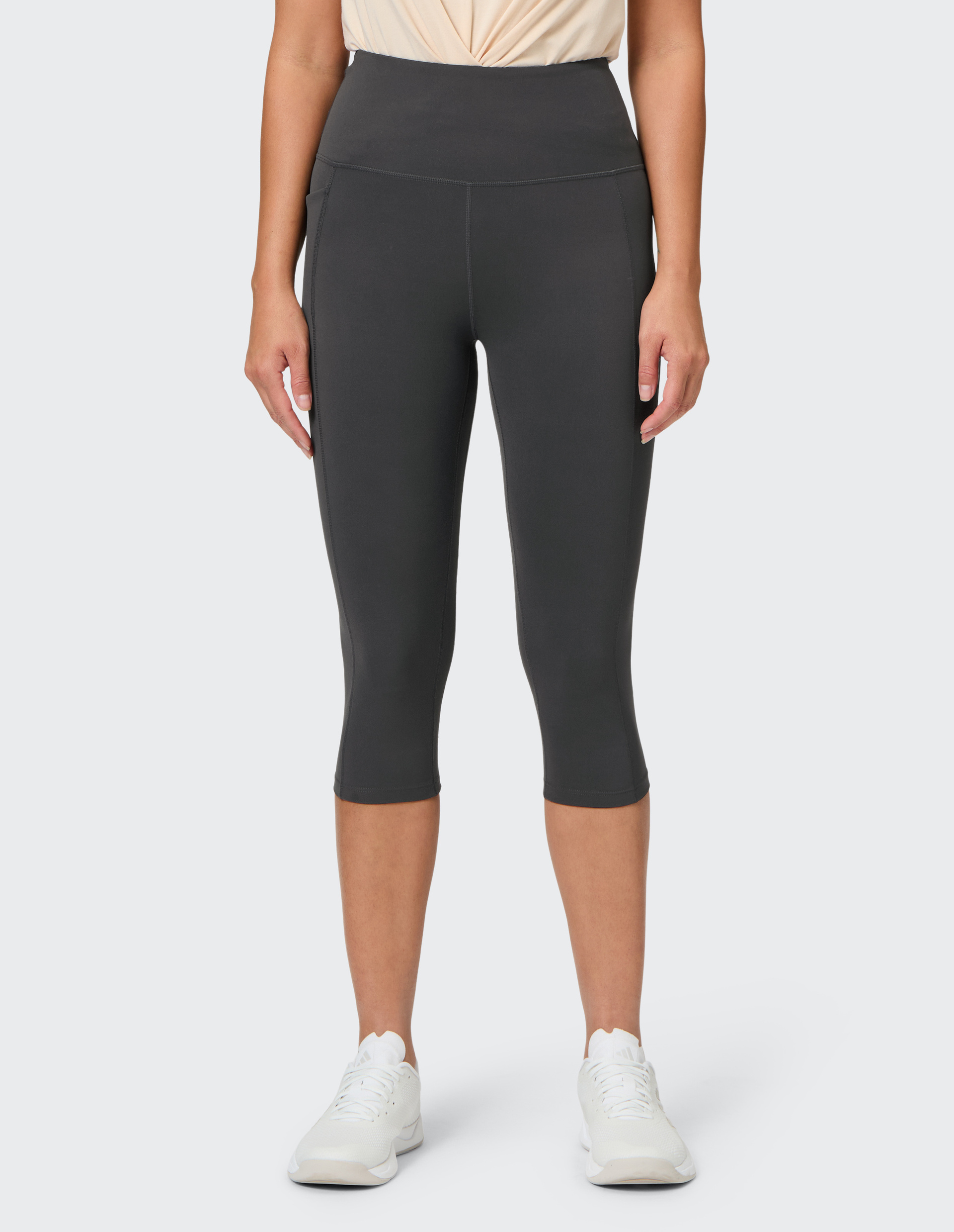 Frau in dunkler Caprihose und weißem Shirt steht frontal, trägt weiße Turnschuhe und zeigt einen sportlichen, lässigen Look.