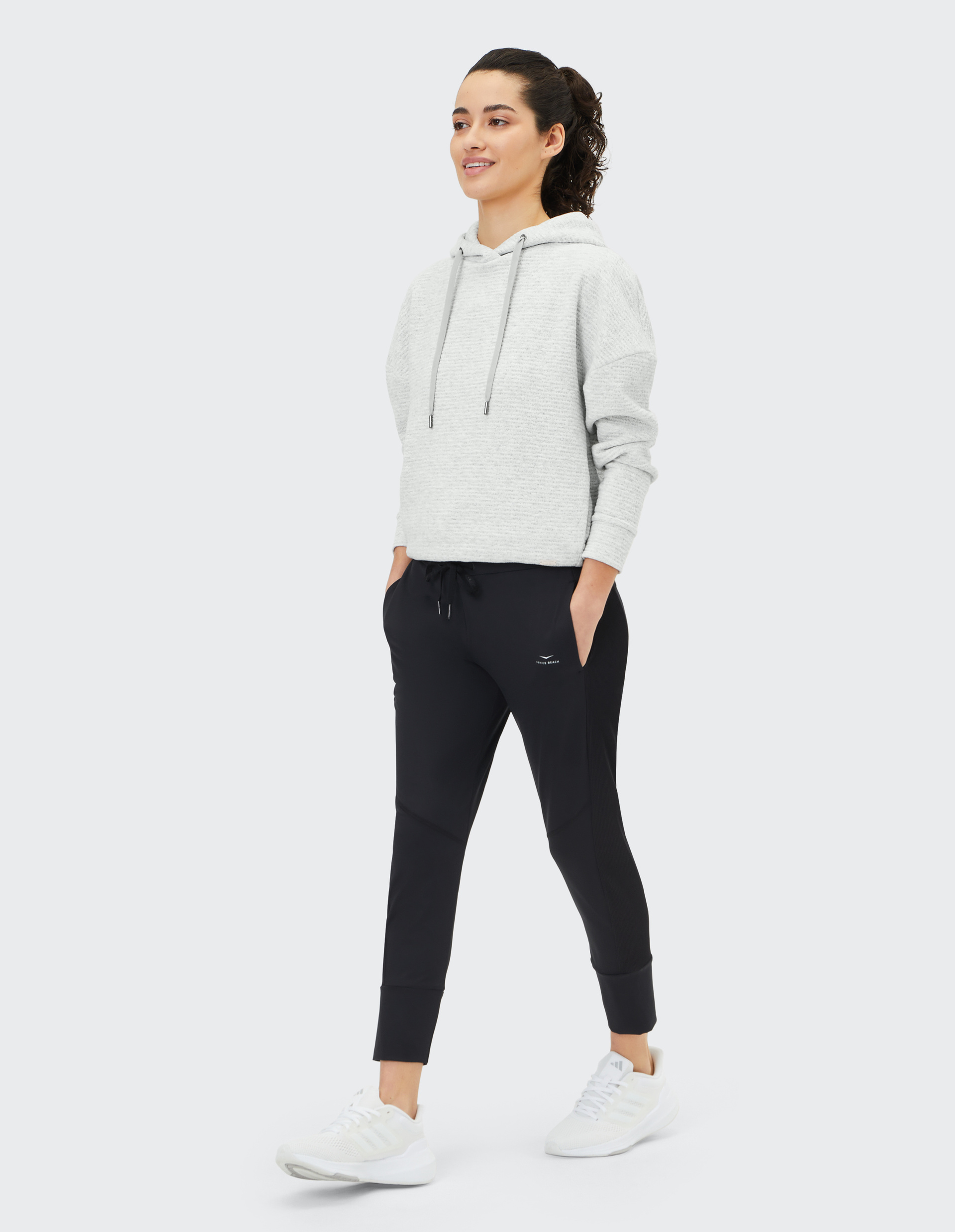 Frau in grauem Hoodie und schwarzer Jogginghose, lächelnd, mit Händen in den Hosentaschen, trägt weiße Sneaker, geht vor weißem Hintergrund.